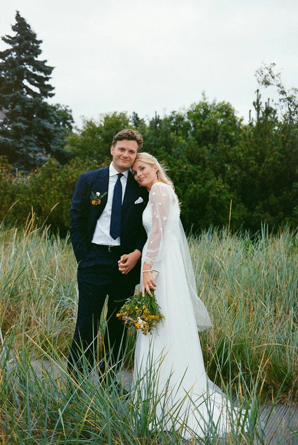 A bride and groom standing in tall grass outdoors, holding hands, with trees in the background. The bride is wearing a white wedding dress and holding a bouquet of yellow and green flowers. The groom is dressed in a dark suit with a white shirt and b