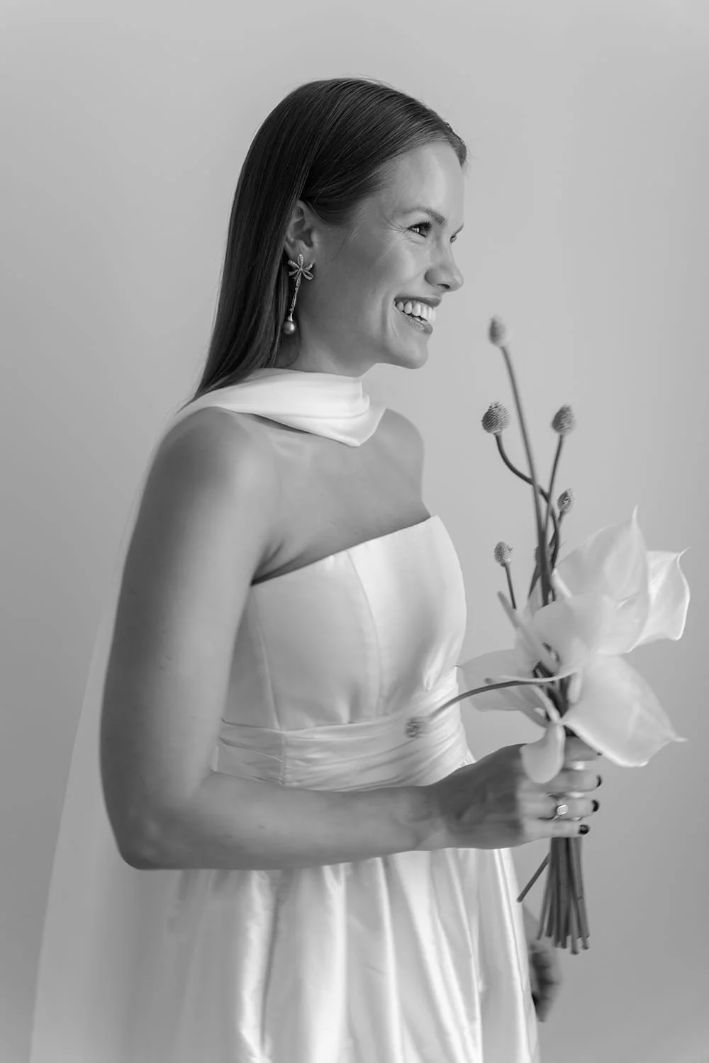 A woman in a strapless satin dress, holding a bouquet of large flowers and small buds, smiling in profile against a plain background.