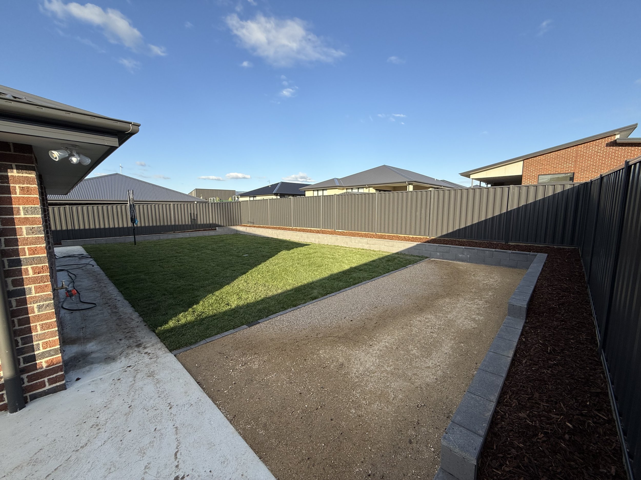 Residential backyard with grass, concrete path, and fenced area under a blue sky.