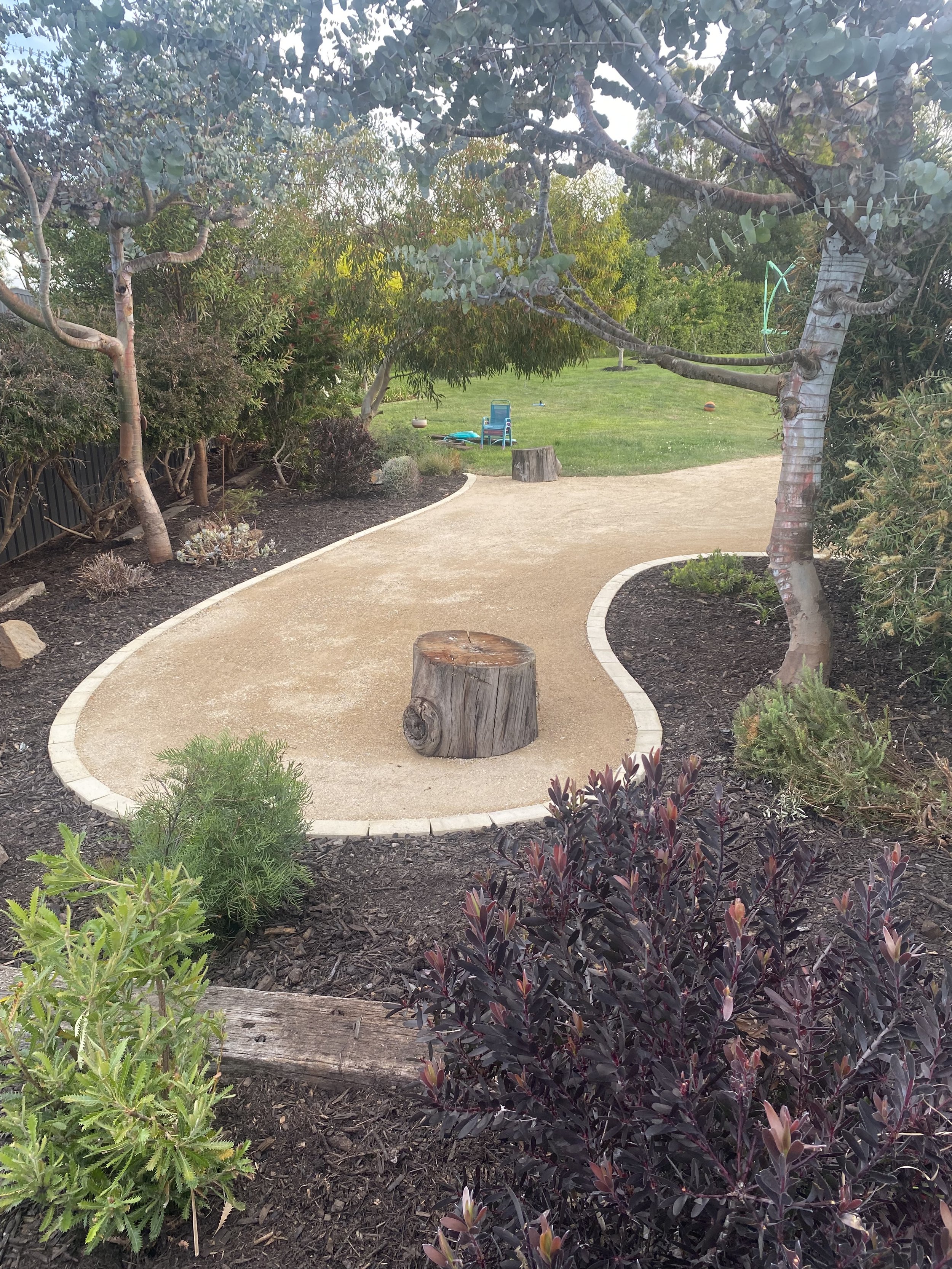 Backyard garden with pathway, trees, shrubs, and a tree stump in the foreground. The path is bordered with small bricks. Lawn area with a garden chair in the background.