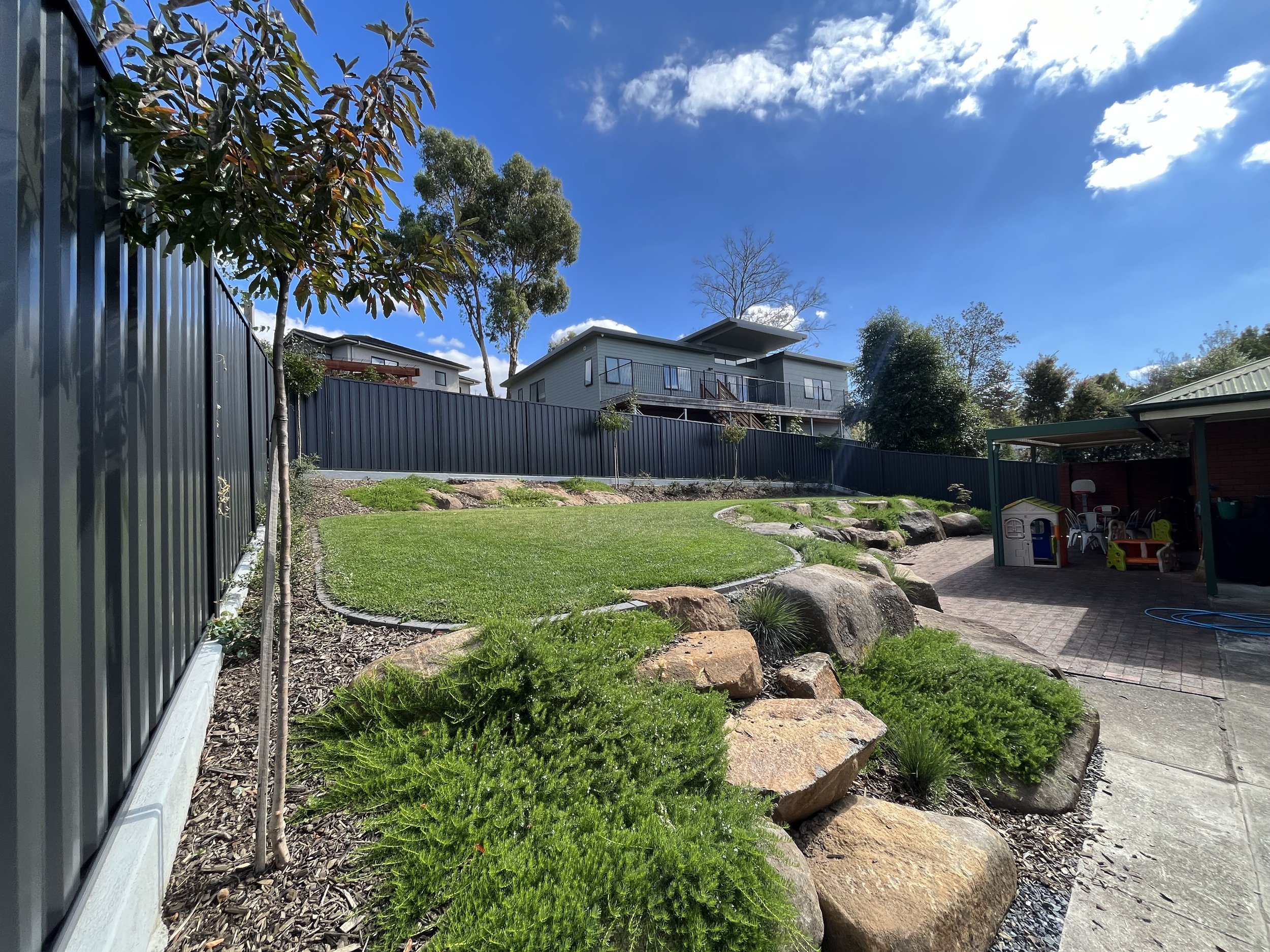 Backyard garden with grass, trees, rocks, and children's playhouse, under blue sky.