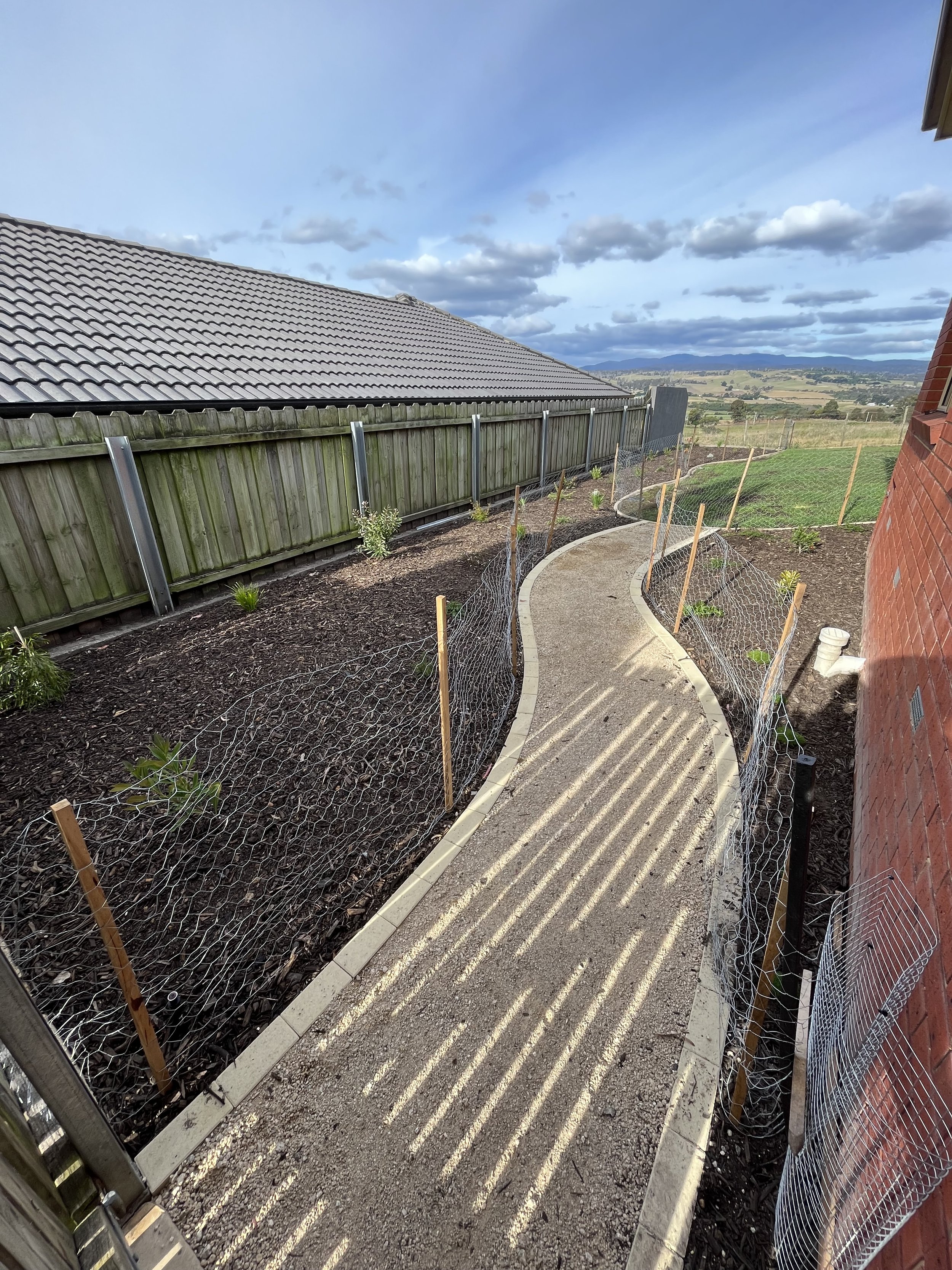 Curved garden pathway with wire fencing, adjacent to a wooden fence and brick wall, under a cloudy sky.