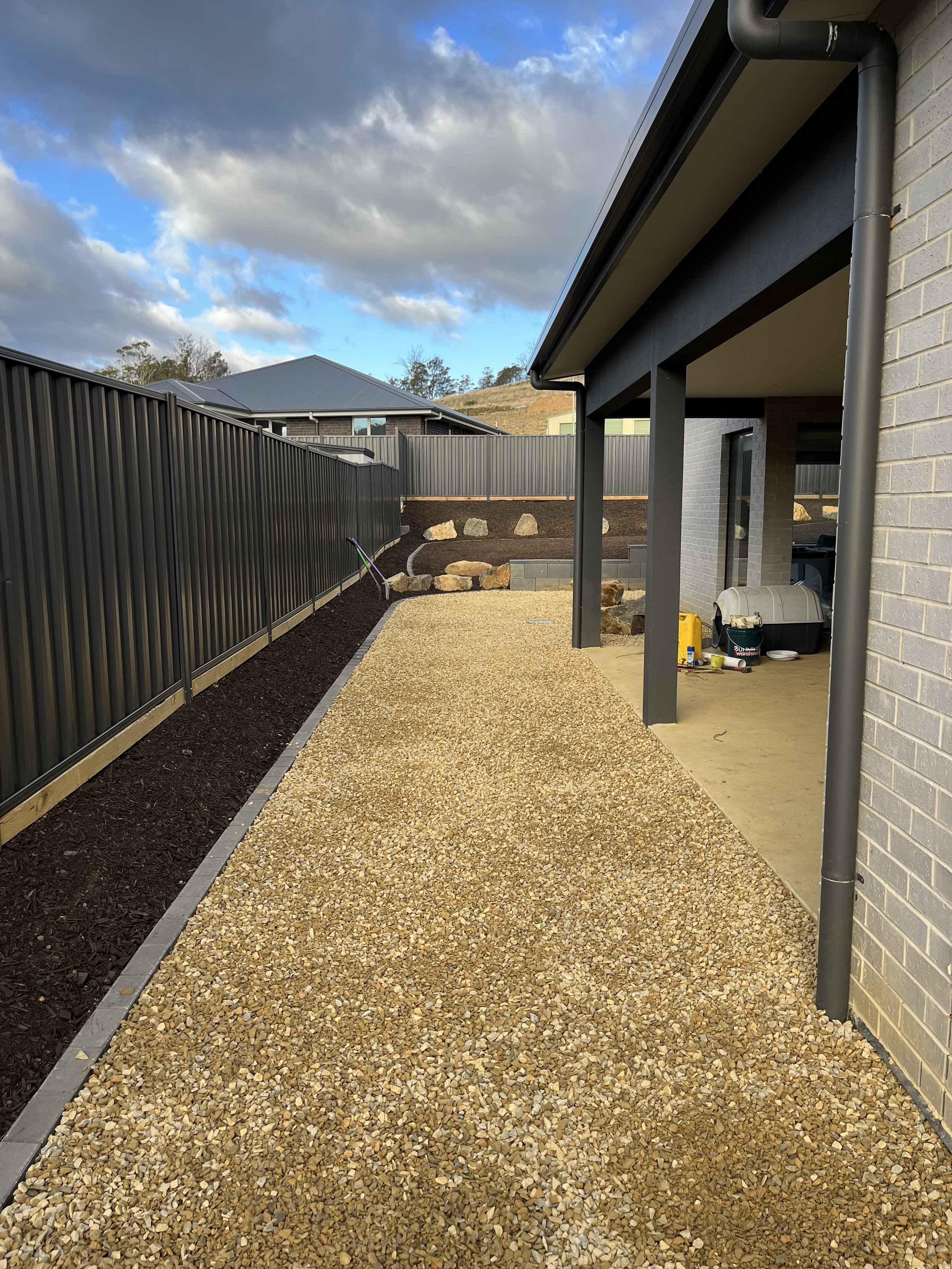 Backyard with gravel path beside a house, metal fence, and cloudy sky.