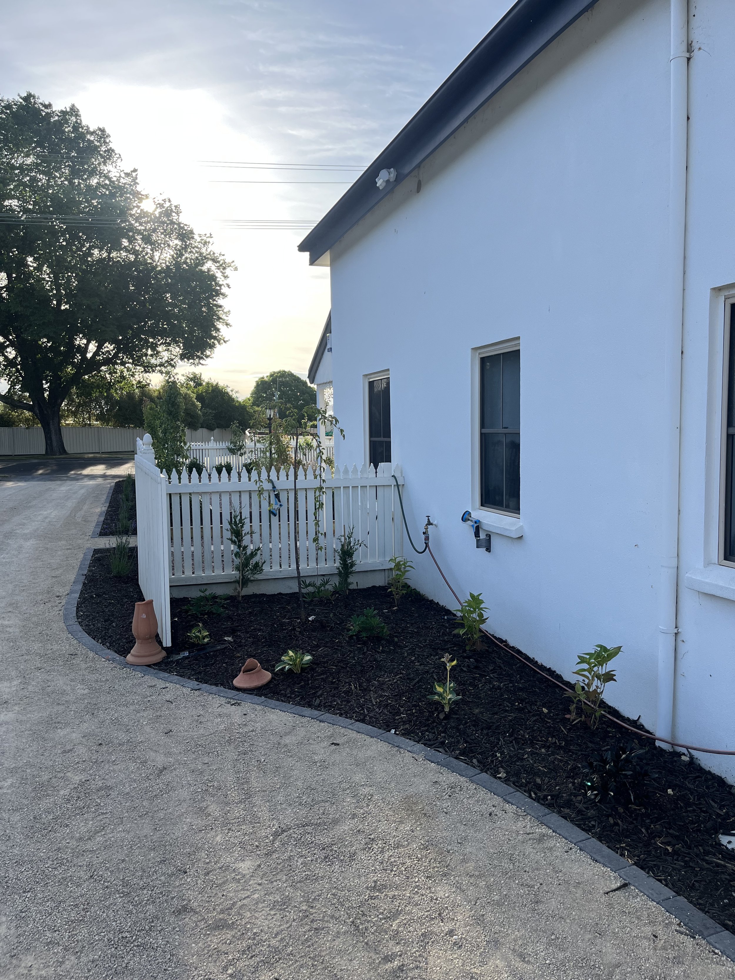 White house exterior with a garden bed, mulch, and young plants beside a curved gravel path. A white picket fence and large tree in the background.