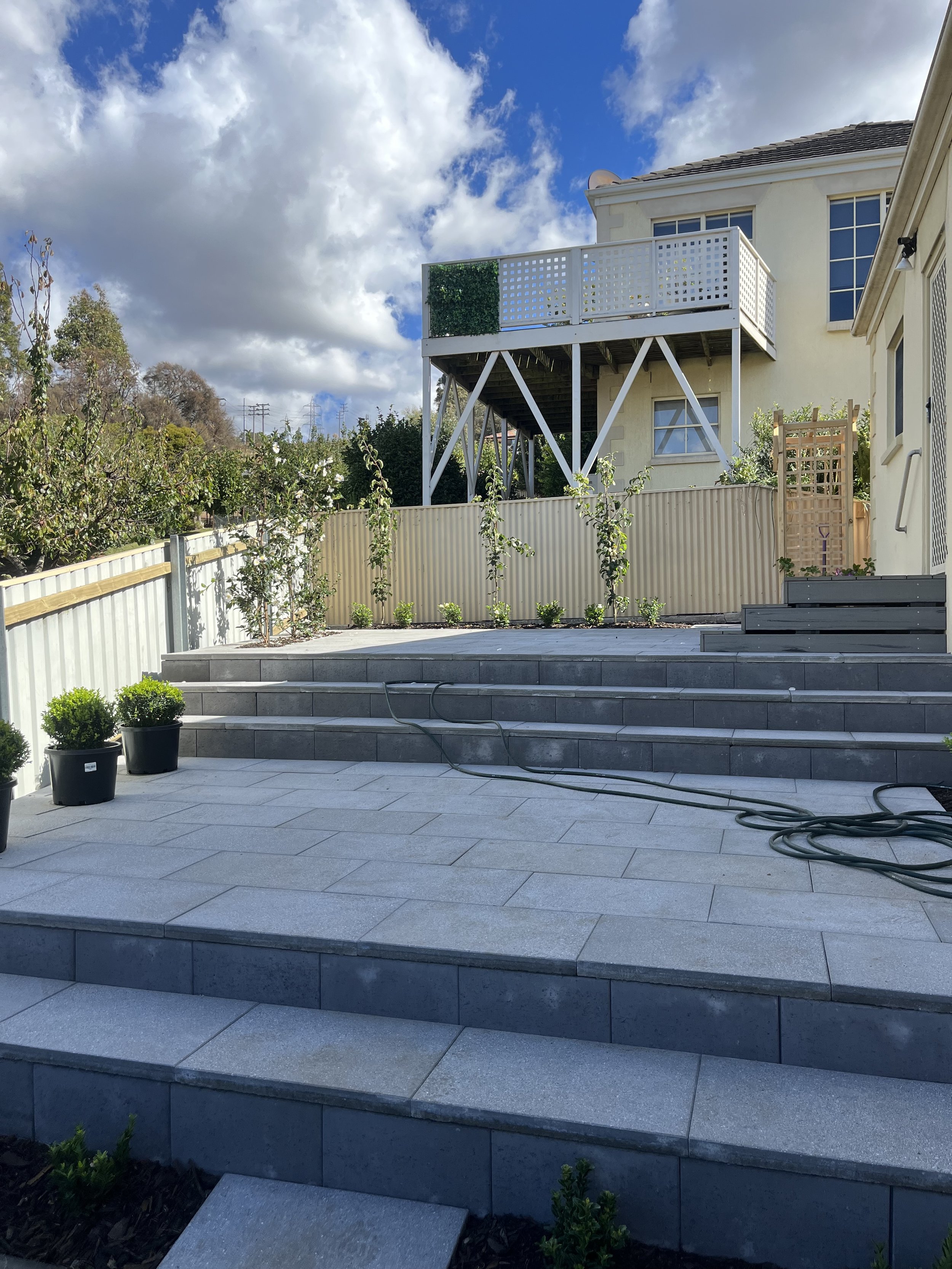 Residential outdoor patio with stairs, potted plants, and a balcony in the background. Bright day with clouds in the sky.