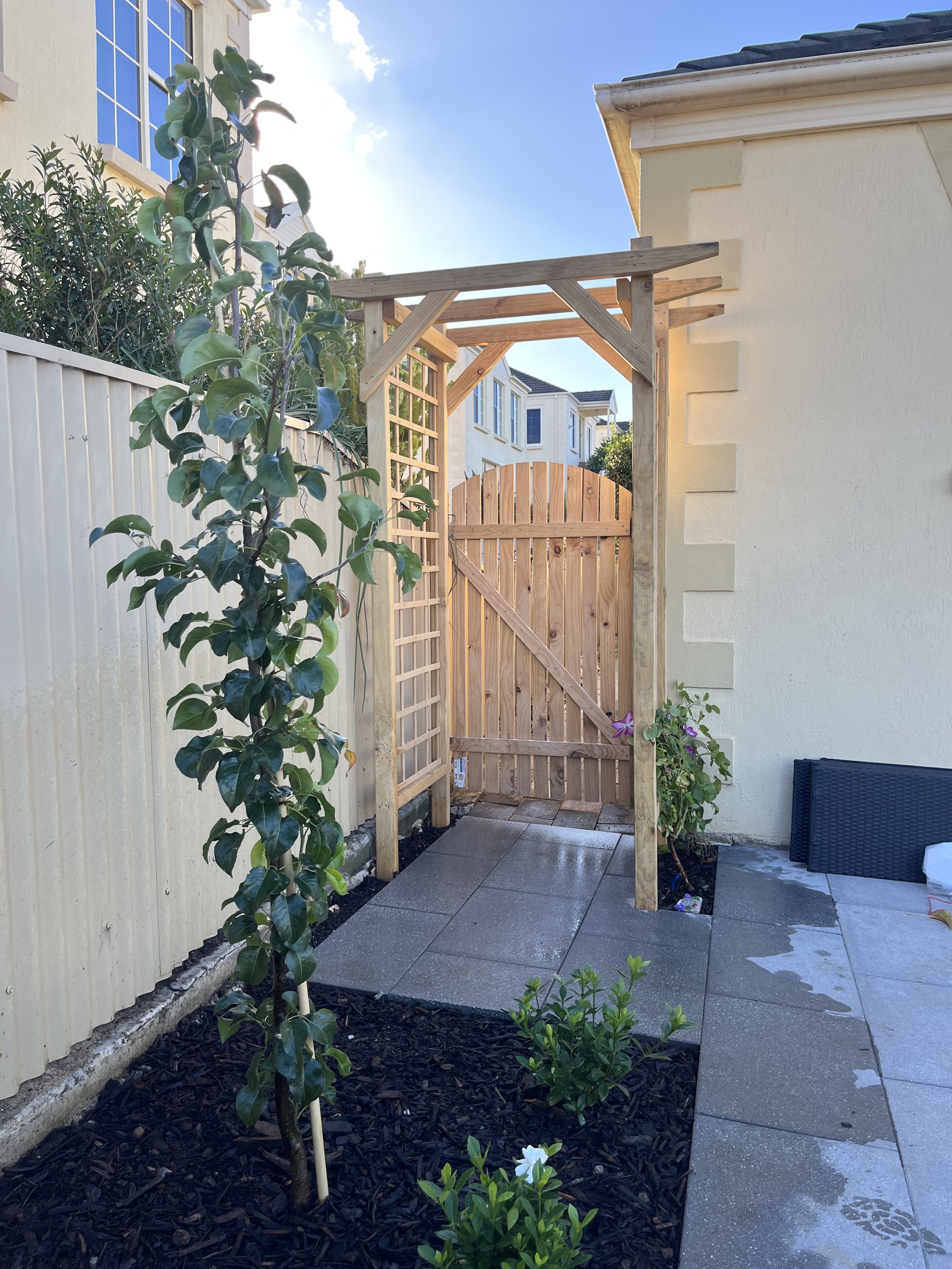 Outdoor garden pathway with a wooden archway and gate, alongside plants and a small tree, bordered by a light-colored building and fence.