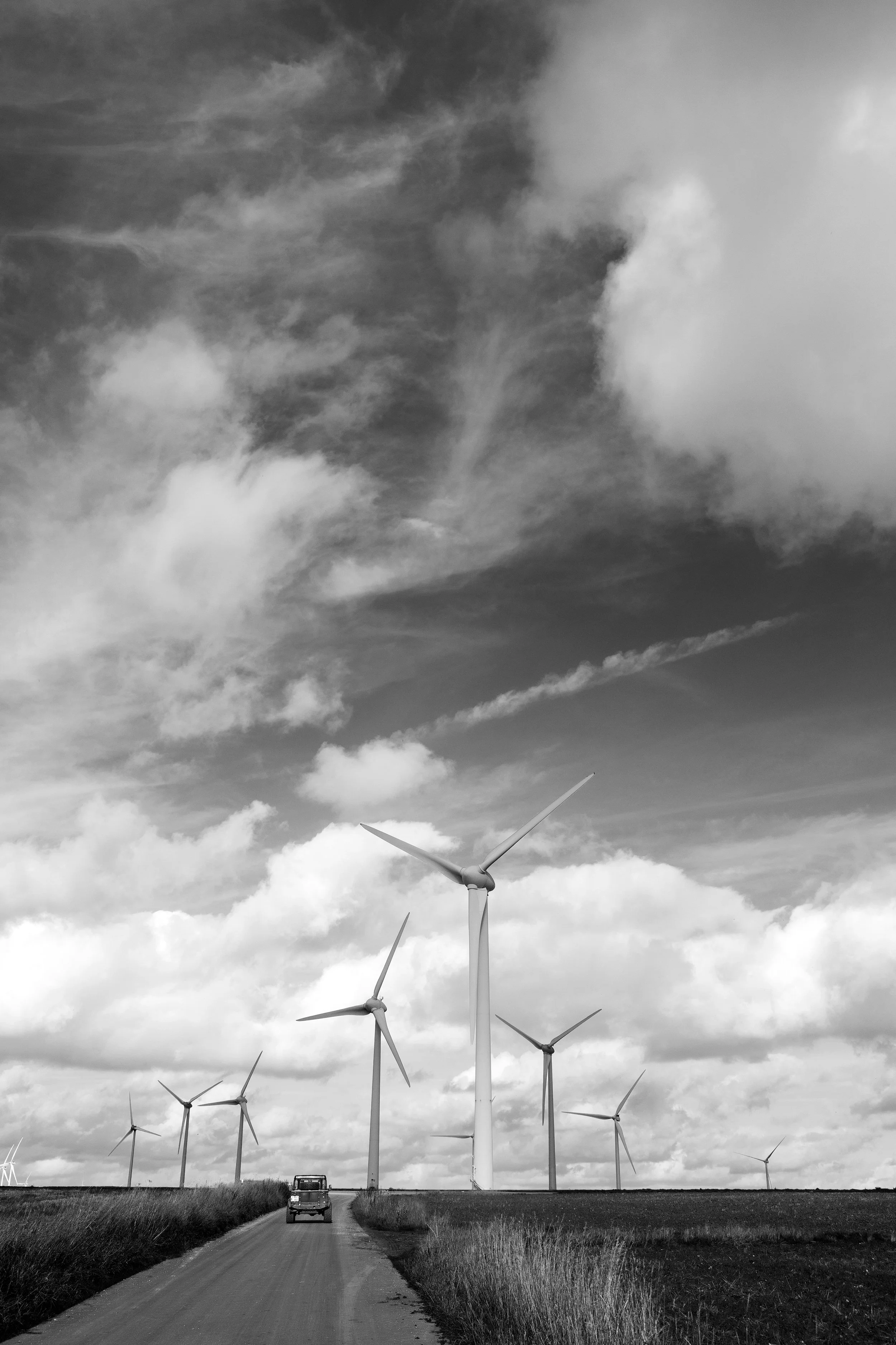 Black and white photo of a rural road leading to wind turbines in a field, under a cloudy sky.
