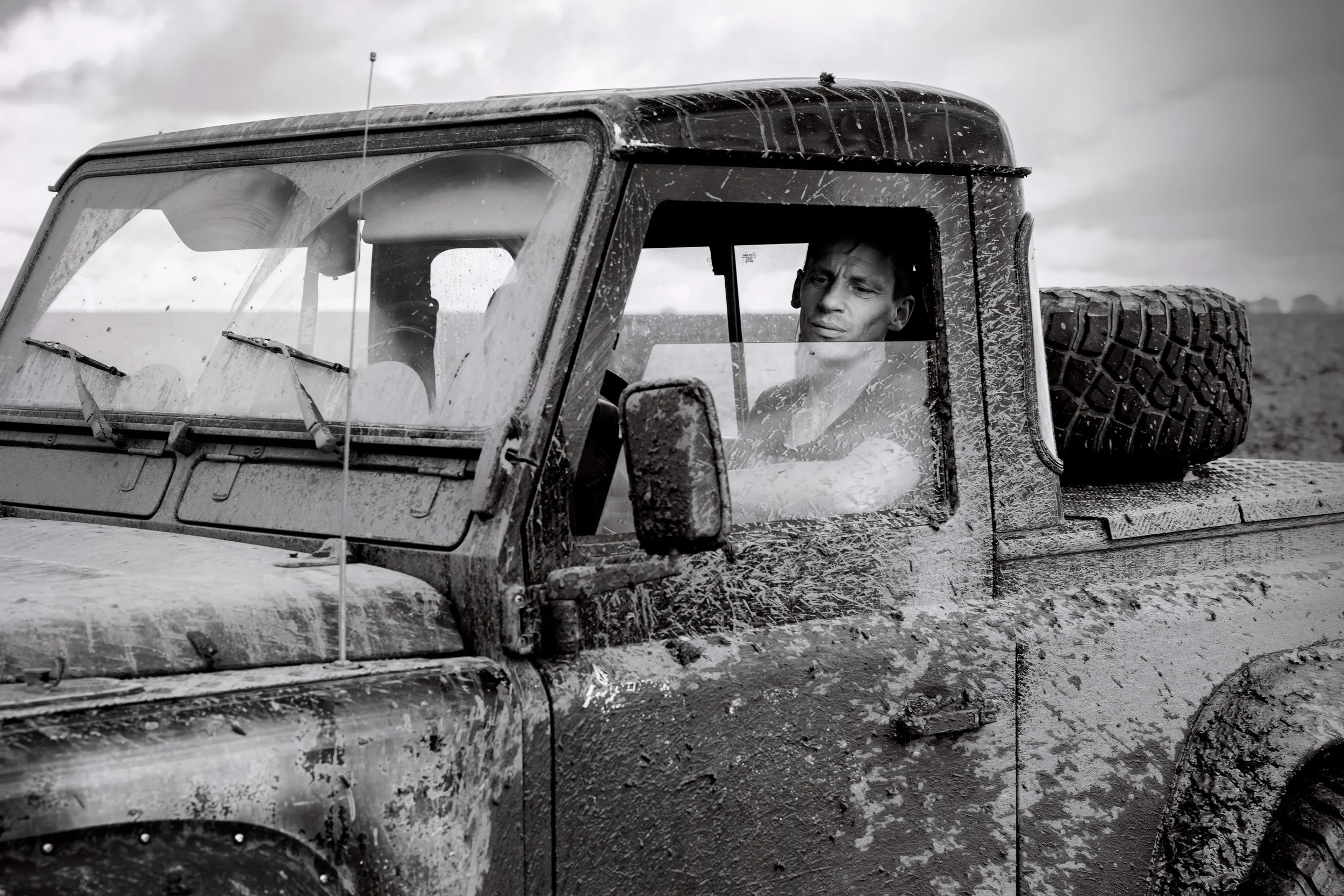 A man sitting in a muddy, dusty off-road vehicle with a spare tire mounted on the back, looking out the window with a serious expression in an outdoor setting.