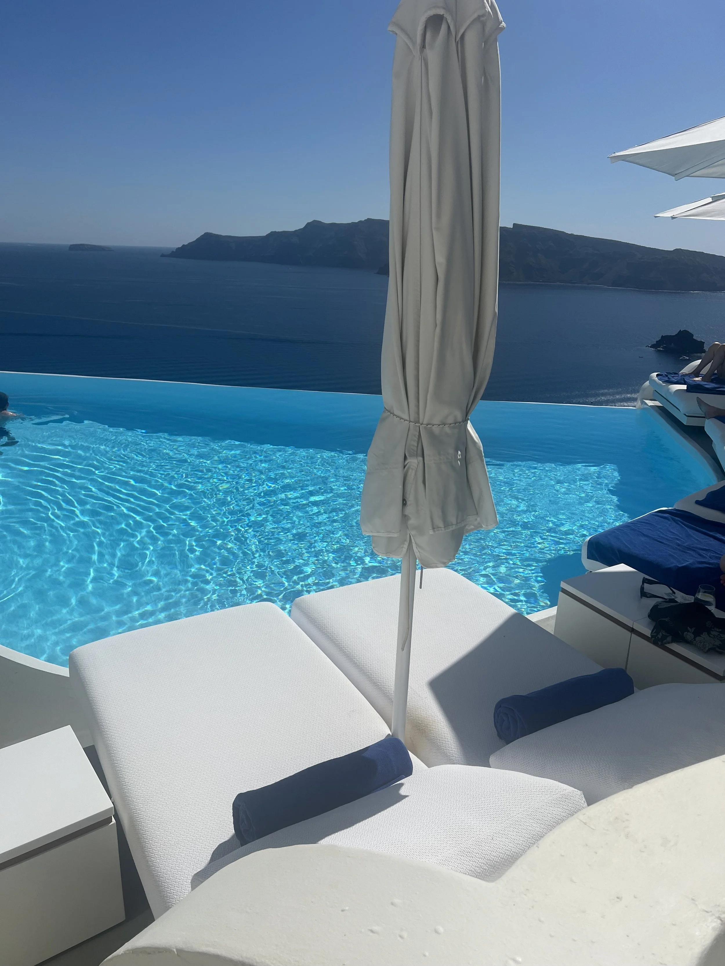 Poolside lounge chairs at a luxury hotel overlooking a panoramic view of Santorini, Greece, personally scouted by Travel by Angela