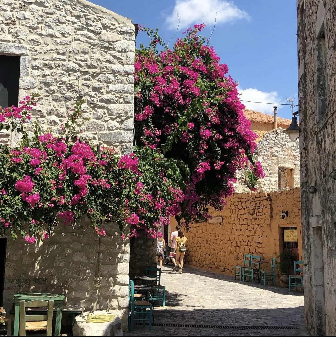 A cobblestone street in Mani, Peloponnese with stone buildings and vibrant pink bougainvillaea flowers climbing the walls, recommended for Travel by Angela custom itineraries
