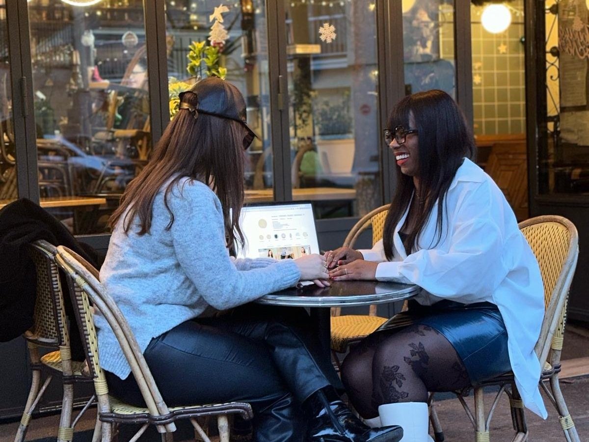 Deux femmes assises à une table de café, discutant et riant. L'une porte un pull gris, un pantalon en cuir noir et une casquette, l'autre porte une blouse blanche, une jupe en cuir noir, des collants noirs et des bottes blanches. Elles utilisent un ordinateur portable.