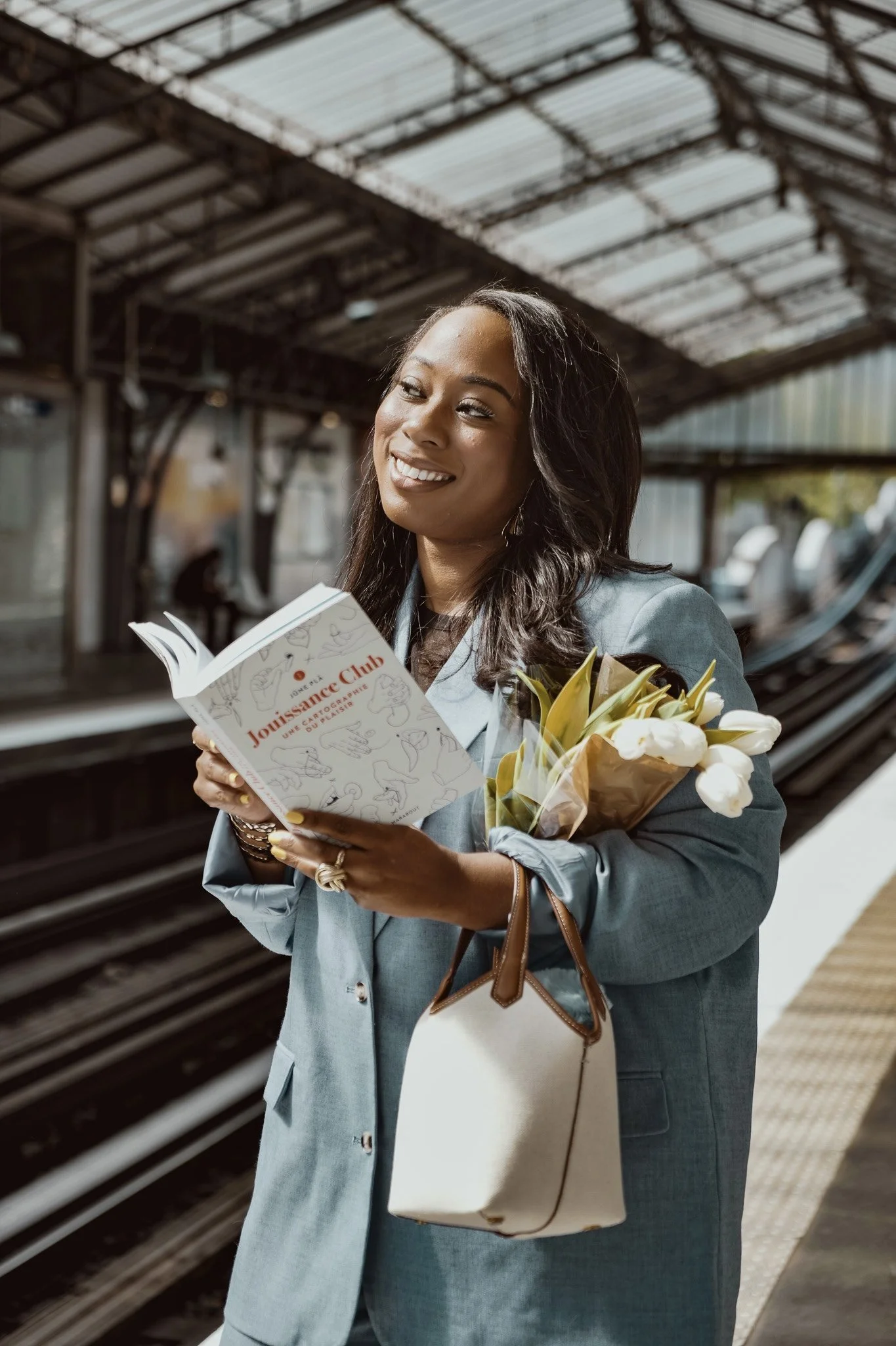 Femme souriante dans une gare, tenant un livre intitulé « Jounaisse Club », un bouquet de fleurs dans l'autre bras, portant un sac blanc avec des détails marron.