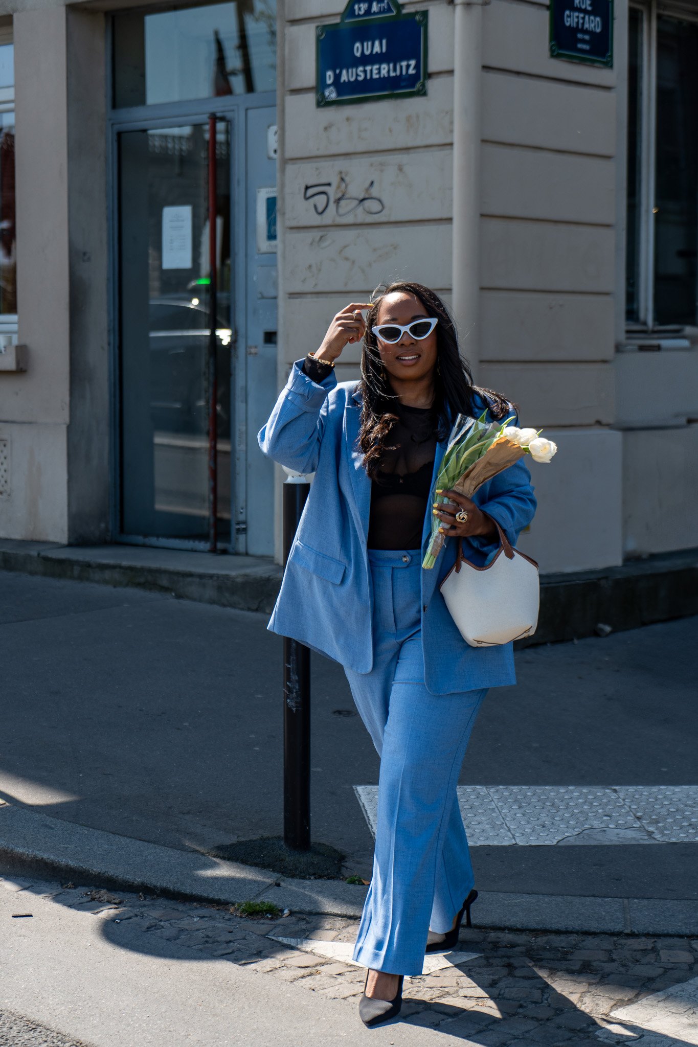 Une femme élégante portant un costume bleu, des lunettes blanches et tenant un bouquet de fleurs marche dans la rue à Paris, près de la rue Giffard et du quai d'Austerlitz.