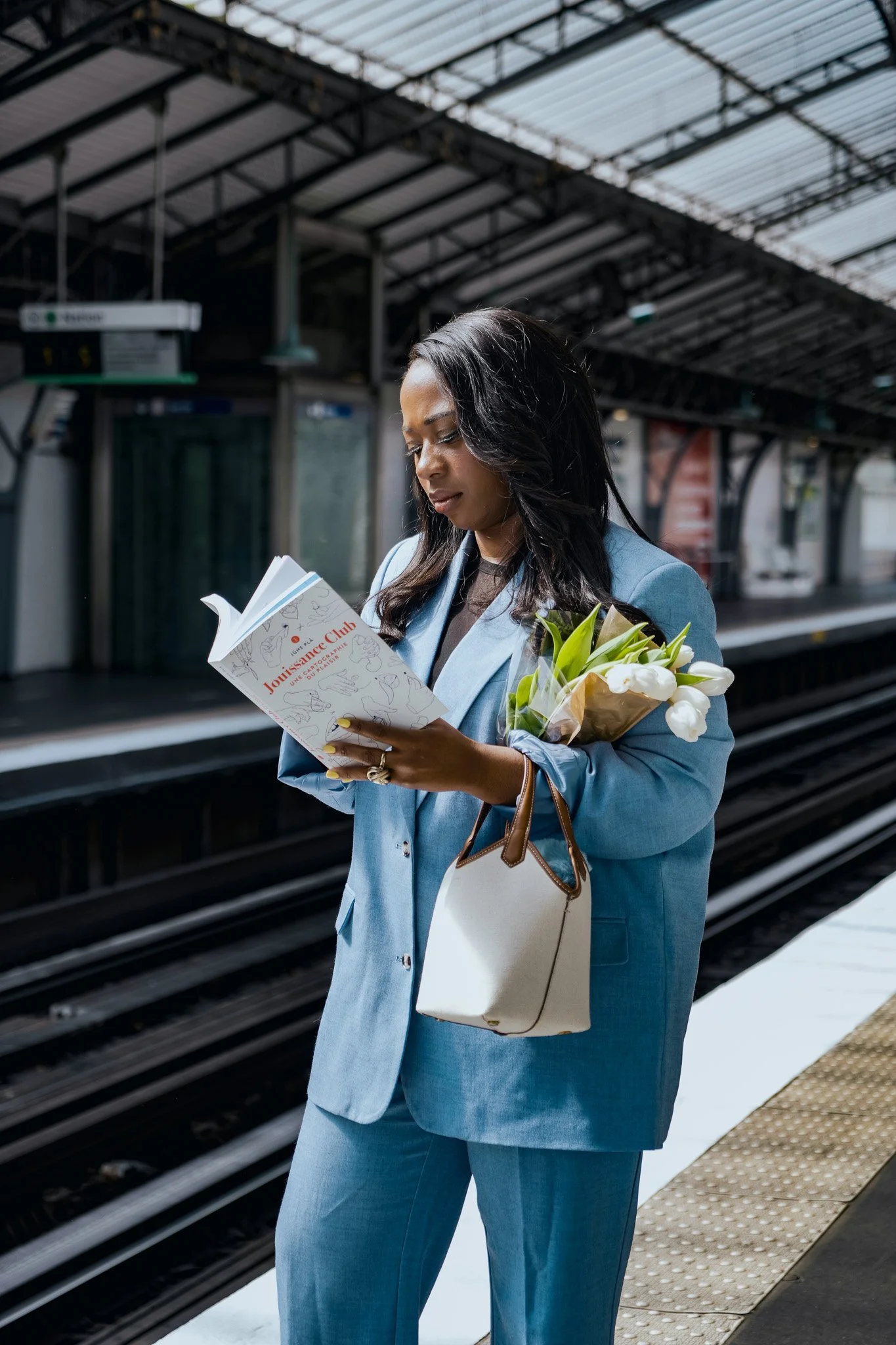 Femme en costume bleu lisant un livre à la station de métro, tenant un bouquet de fleurs et un sac blanc.