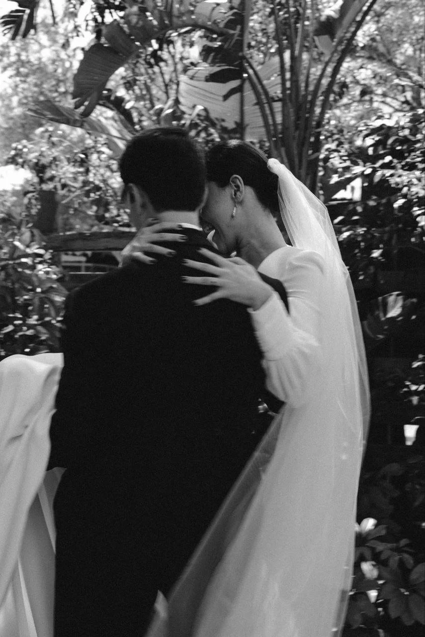 A black and white photo of a bride and groom embracing, with the bride in a white wedding dress and veil, and the groom in a dark suit, surrounded by lush foliage.