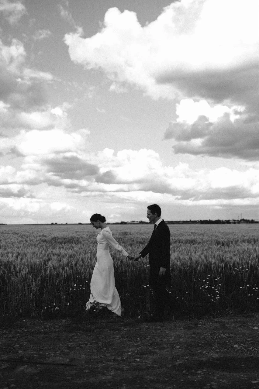 A black and white photo of a couple walking hand in hand through a grassy field under a cloudy sky, with the woman in a long white dress and the man in a suit.