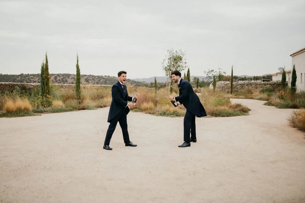 Two men in suits celebrating outdoors, holding champagne bottles and glasses, on a dirt path with cypress trees and desert landscape background.