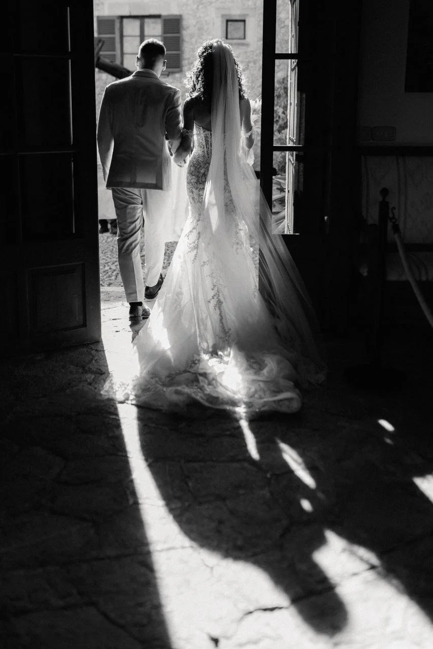 A bride and groom walking out of a building, holding hands, with sunlight casting a shadow of their figures on the ground.