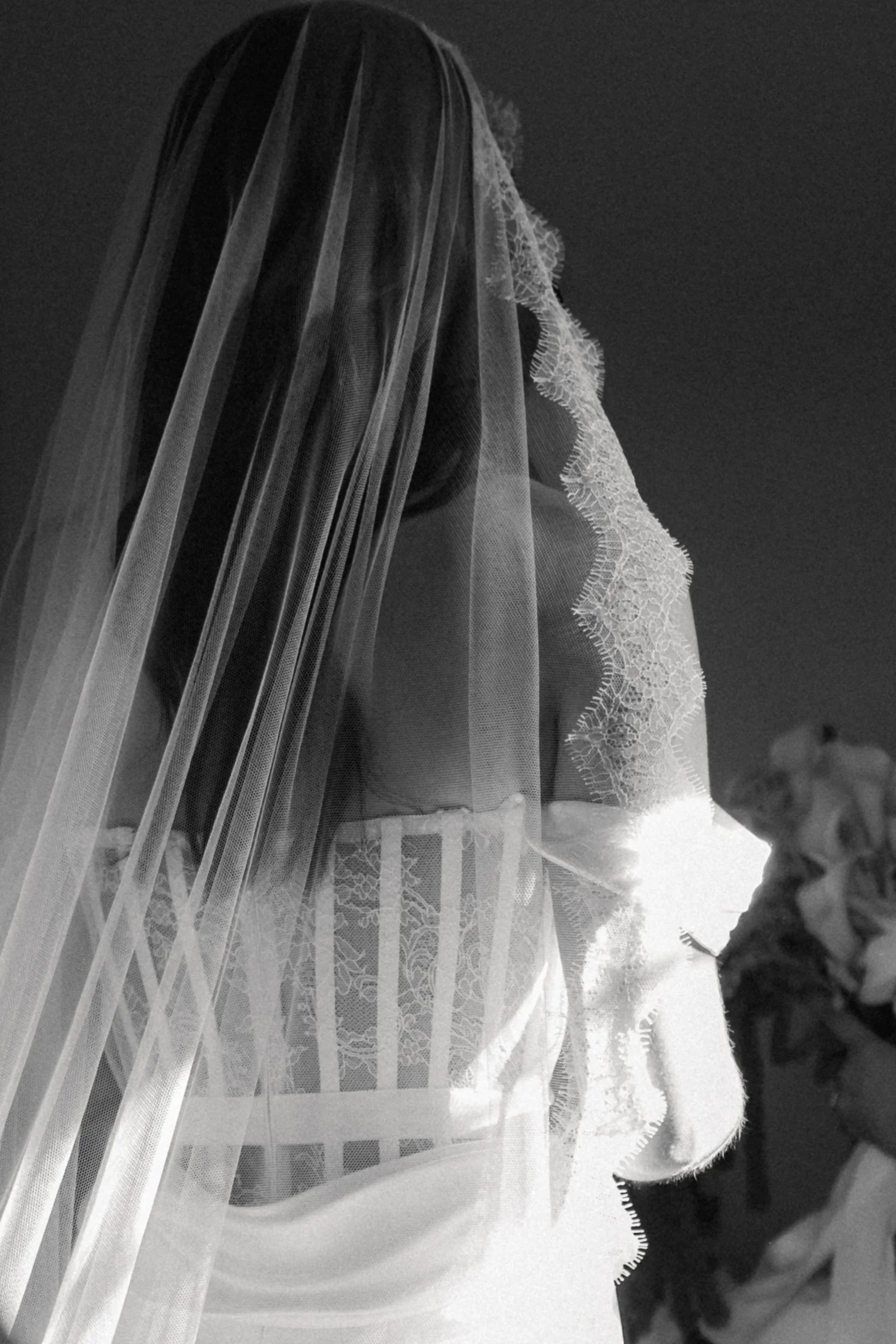 A woman wearing a lace wedding dress and veil, standing outdoors, possibly during a wedding ceremony, with a dark sky and flowers in the background.