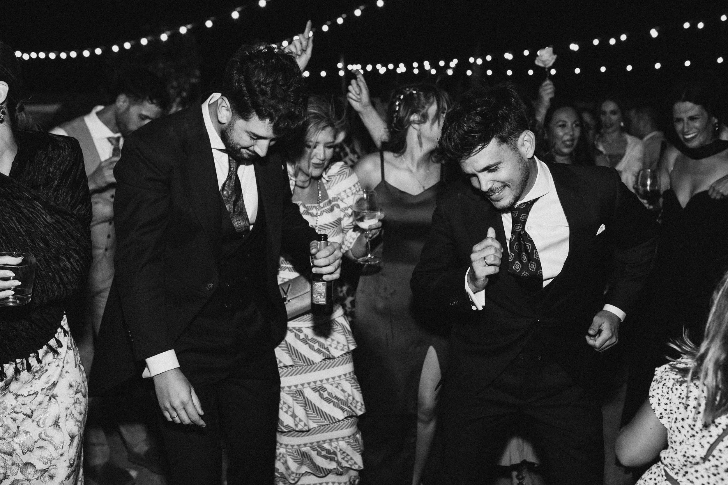 People dancing and socializing at a party with string lights overhead, black and white photo.