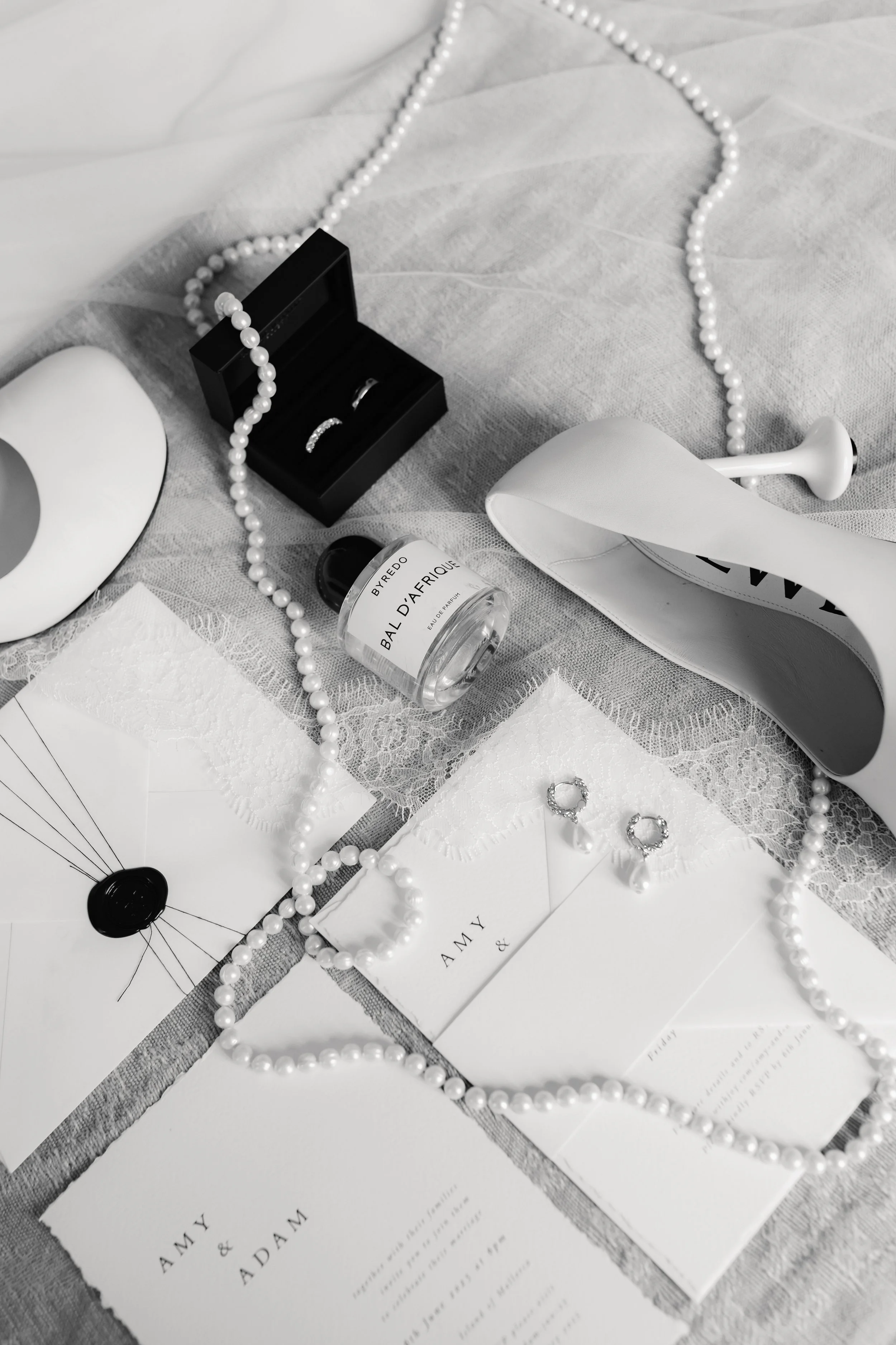 Black jewelry box with rings on a white lace cloth, surrounded by pearl necklaces, white candles, perfume bottle, and wedding invitation.