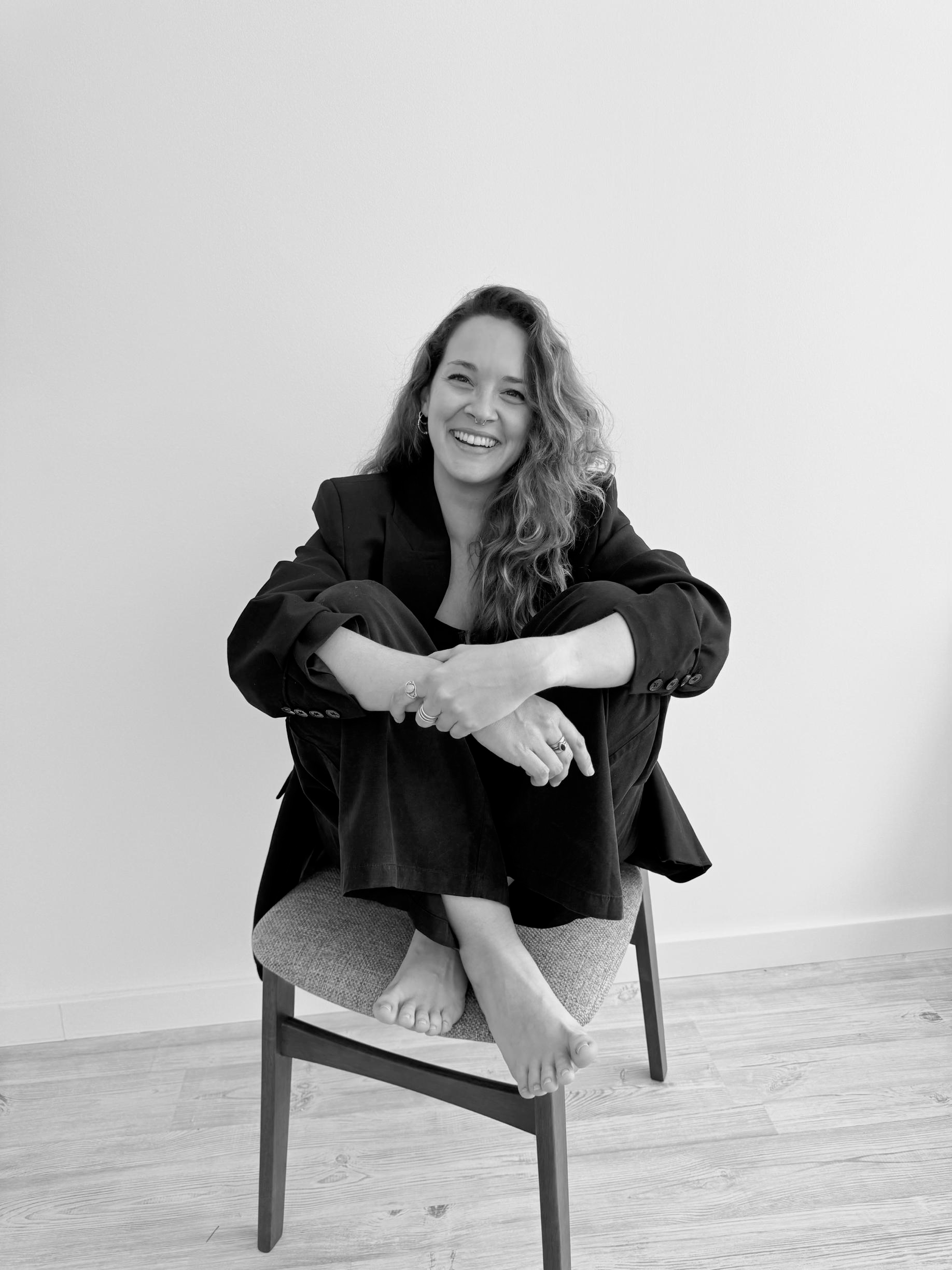 Black and white photo of a woman sitting on a chair with her legs crossed and feet resting on the seat. She is smiling and has long, wavy hair. She is wearing a dark blazer and pants, against a plain wall background.