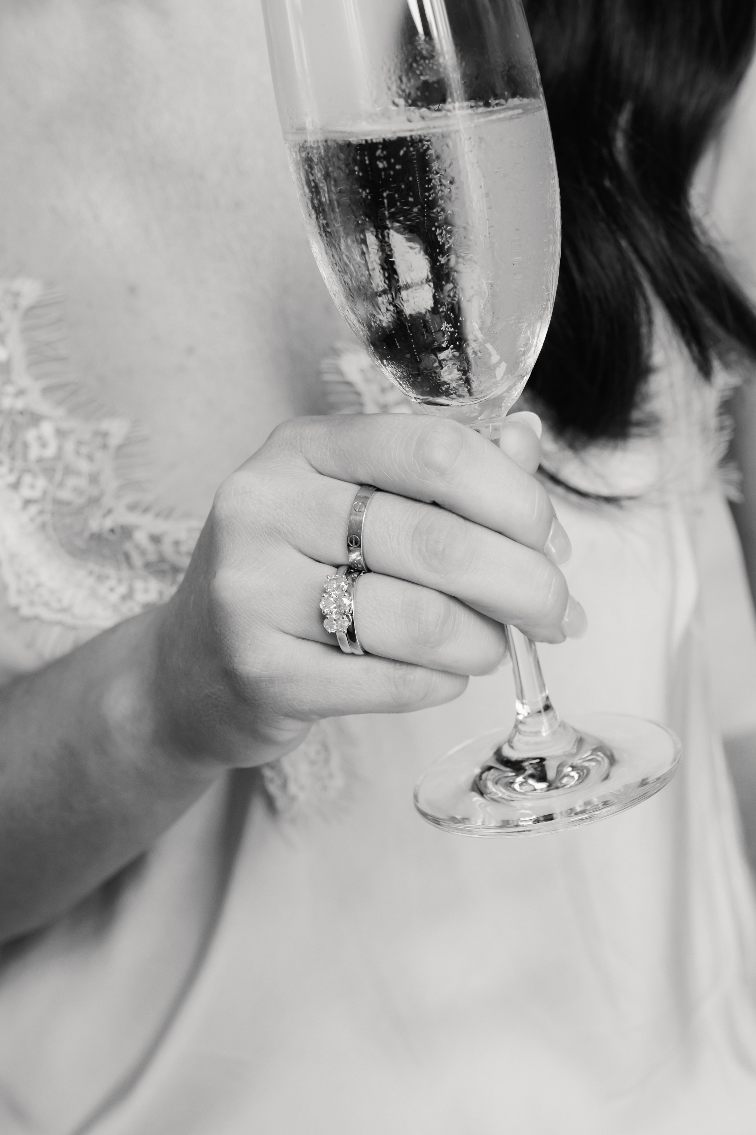 A woman holding a champagne glass filled with sparkling wine, wearing rings including a large engagement ring, with her face partially visible.