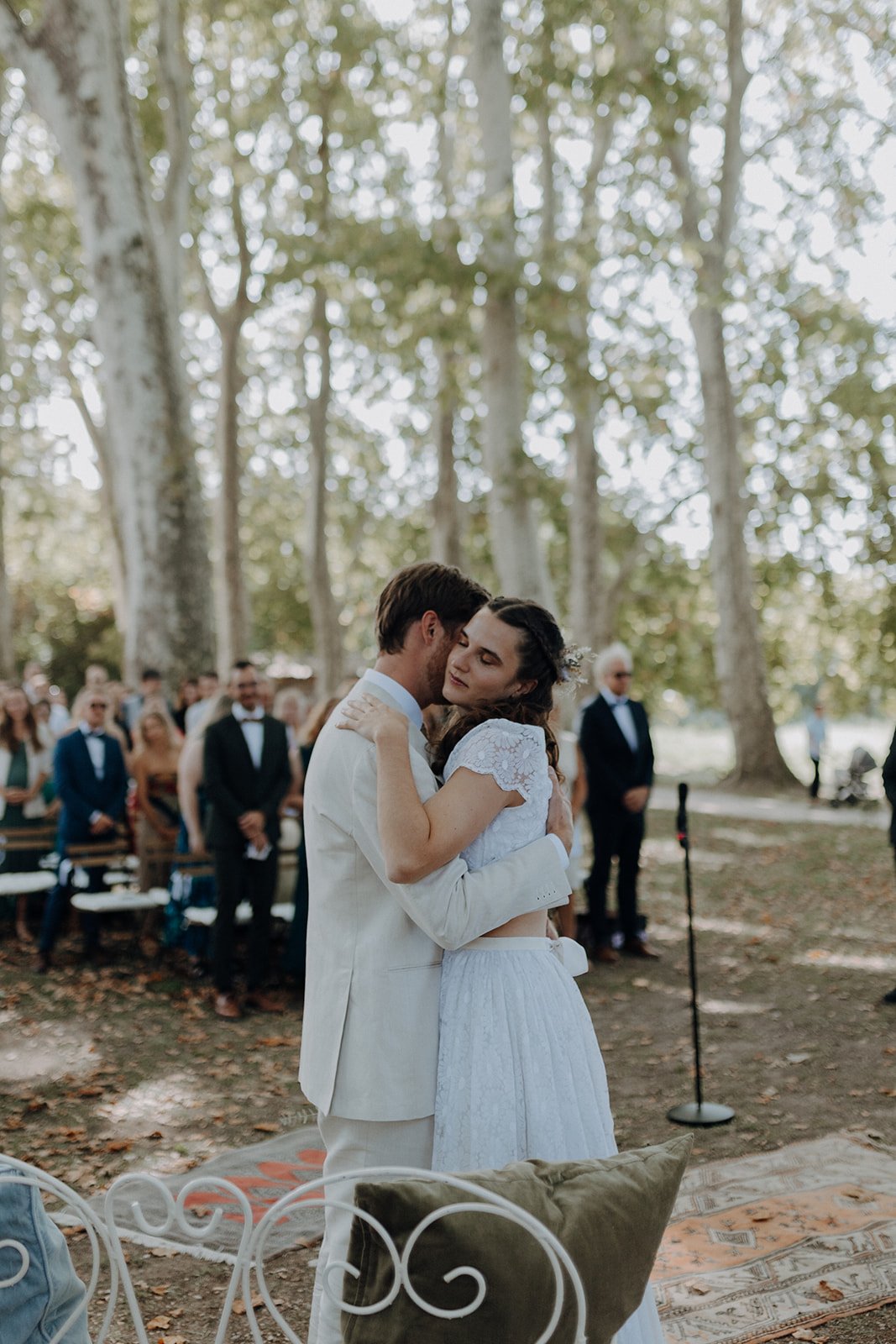 Brautpaar umarmt sich während einer Hochzeit im Freien, umgeben von Bäumen und Hochzeitsgästen.