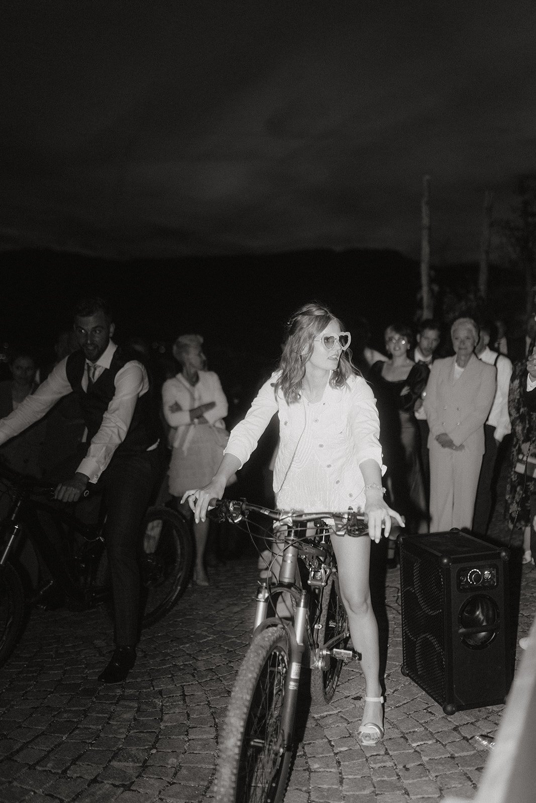 Eine Frau auf einem Fahrrad, umgeben von einer Gruppe von Menschen, bei Nacht. Die Frau trägt Herzbrillen und eine helle Jacke. Neben ihr steht ein Lautsprecher.