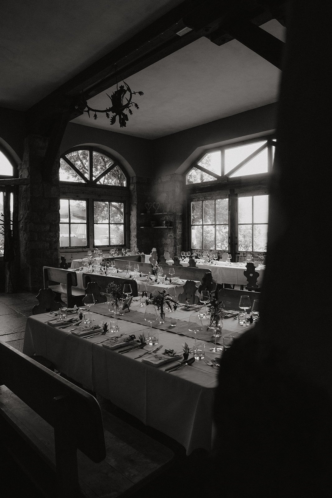 Schwarz-Weiß-Foto eines festlich gedeckten Esszimmers mit langen Tischen, dekoriert mit Kerzen und Blumenarrangements vor großen Fenstern mit Blick nach draußen.