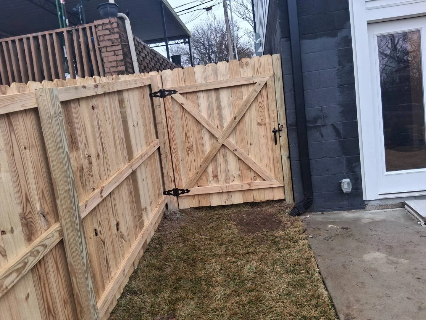 Custom wood fence gate with X-brace design and black iron hinges and latch next to a home