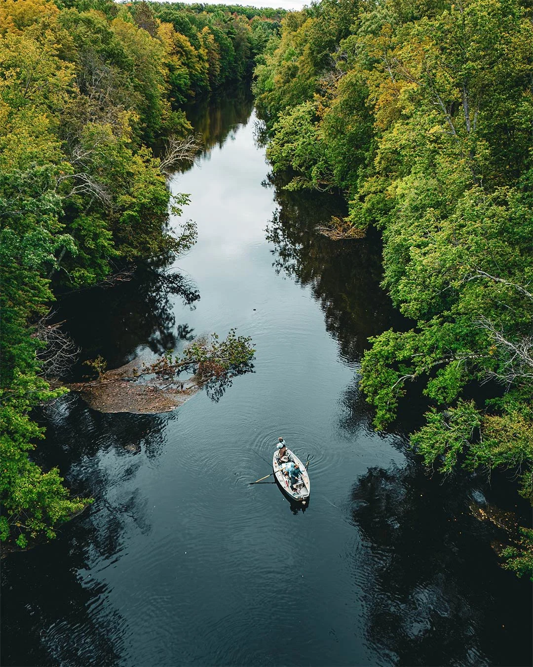 A boat with two people is floating on a narrow river surrounded by dense green trees. The river reflects the trees and the sky above.