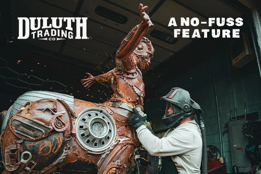 A welder wearing a helmet and gloves welding a large sculpture of a woman riding a motorcycle, made from scrap metal, at Duluth Trading Co. with the caption 'A No-Fuss Feature'.