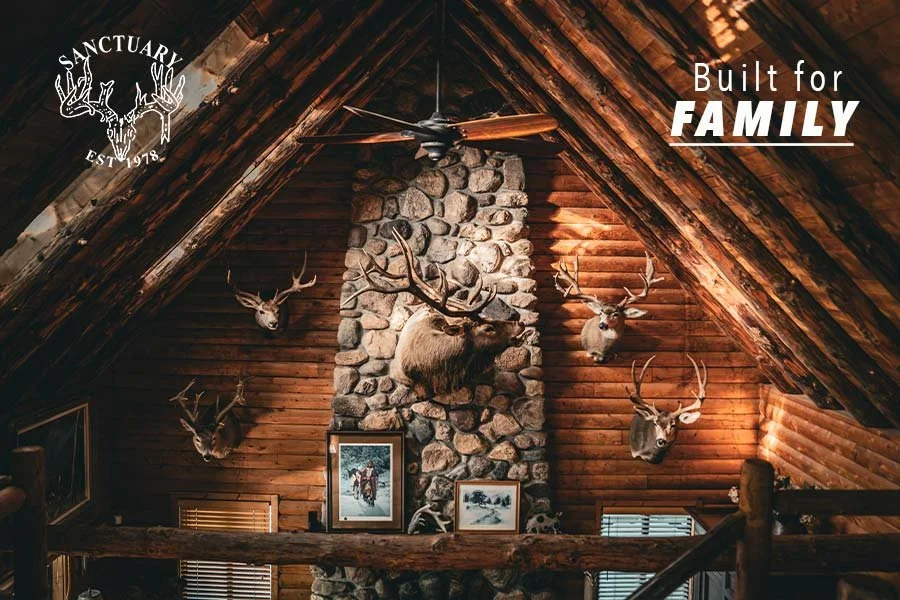 Interior of a rustic log cabin with a stone fireplace, mounted deer heads, wood-paneled walls, and framed pictures. The ceiling has exposed wooden beams and a ceiling fan. The image promotes the message 'Built for Family' and features a Sanctuary logo.