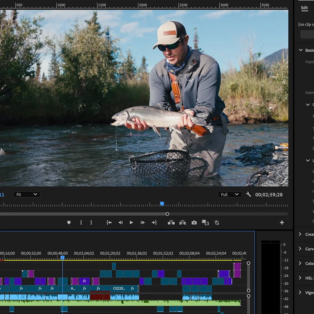 A man wearing a gray jacket, sunglasses, and a beige cap stands in a shallow river, holding a fish with both hands. The background shows trees and a mountain under a partly cloudy sky.