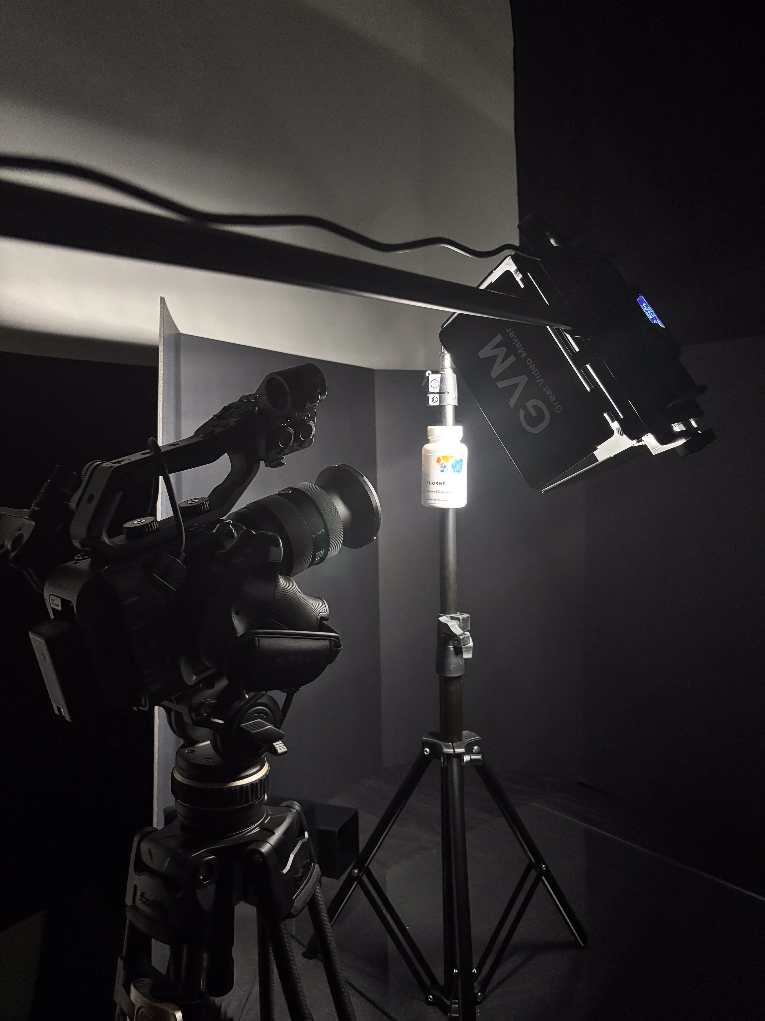 Video production setup with a camera on a tripod, a monitor, and a bottle of artificial tears on a stand, in a dark studio environment.