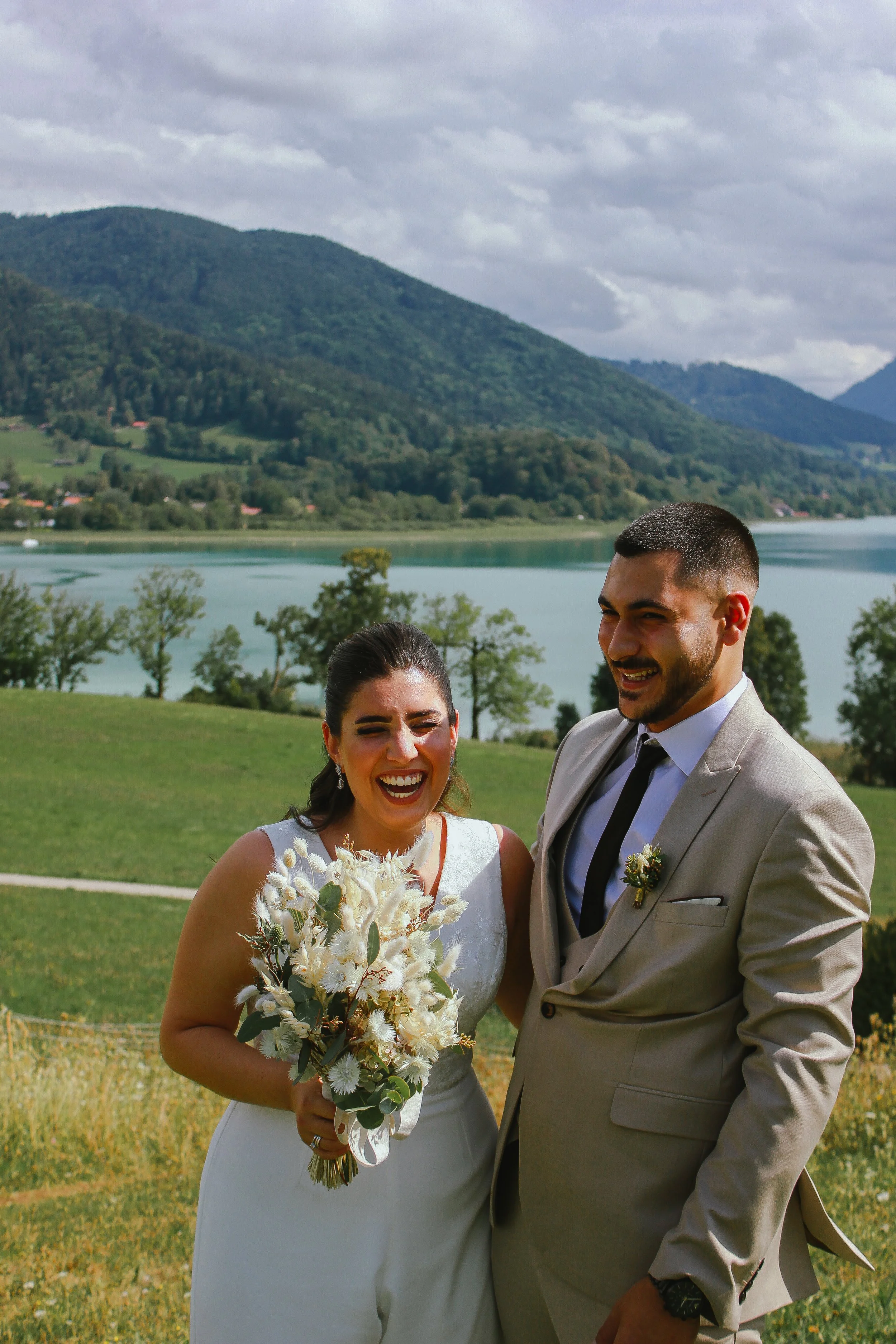 Ein Paar, vermutlich Braut und Bräutigam, lachend im Freien mit Bergen, Wasser und grüner Landschaft im Hintergrund. Aufgenommen von Hilal Studios.