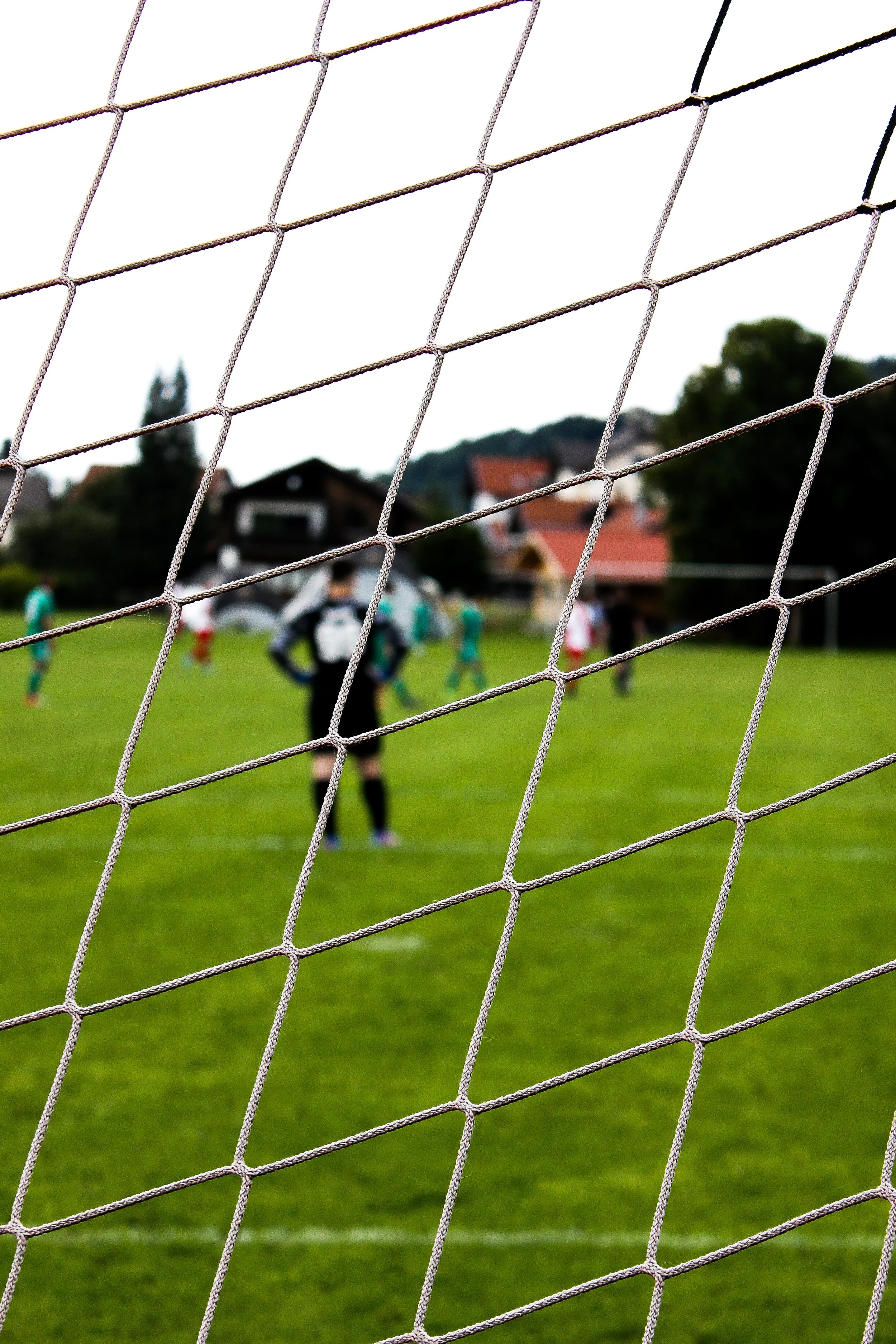 Fußballtor mit einem Spiel im Hintergrund, Spieler auf dem Spielfeld, Häuser im Hintergrund, Blick durch das Netz. Aufgenommen von Hilal Studios.