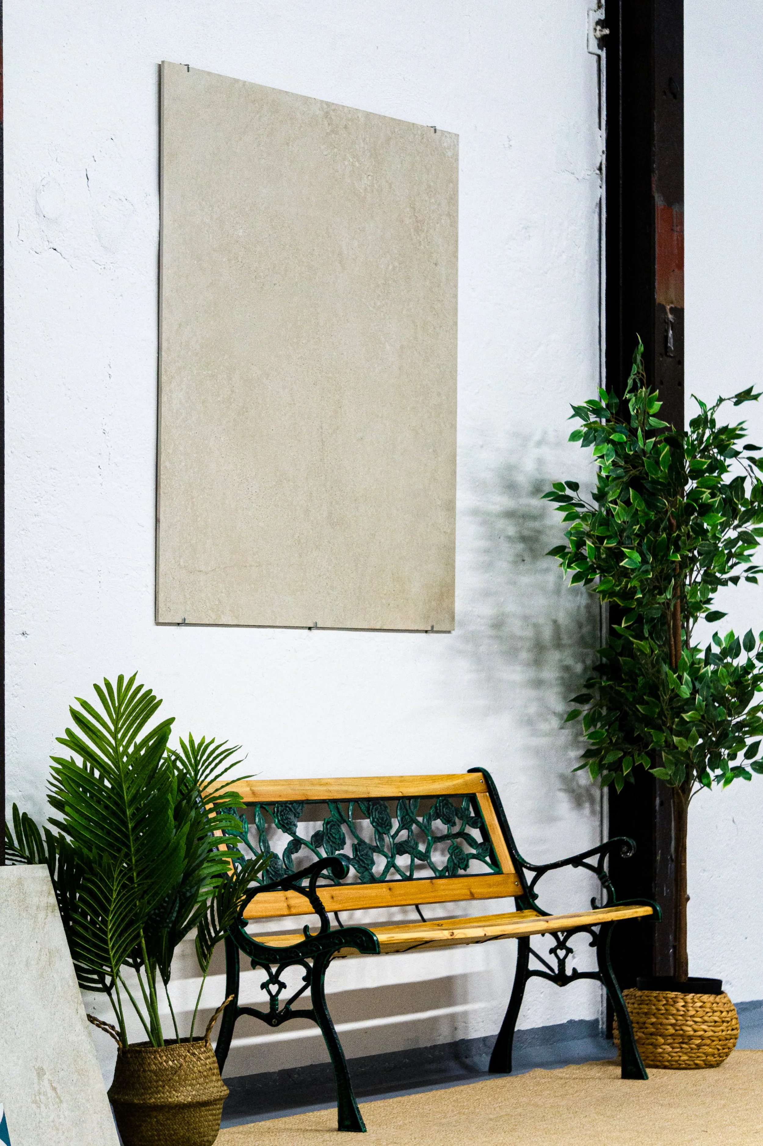 Ein Sitzbank mit schwarzen verzweigten Metallarmen, hölzernen Sitztflächen und Rückenlehne, umgeben von Pflanzen in Körben, vor einer weißen Wand mit einem großen leeren Rahmen.