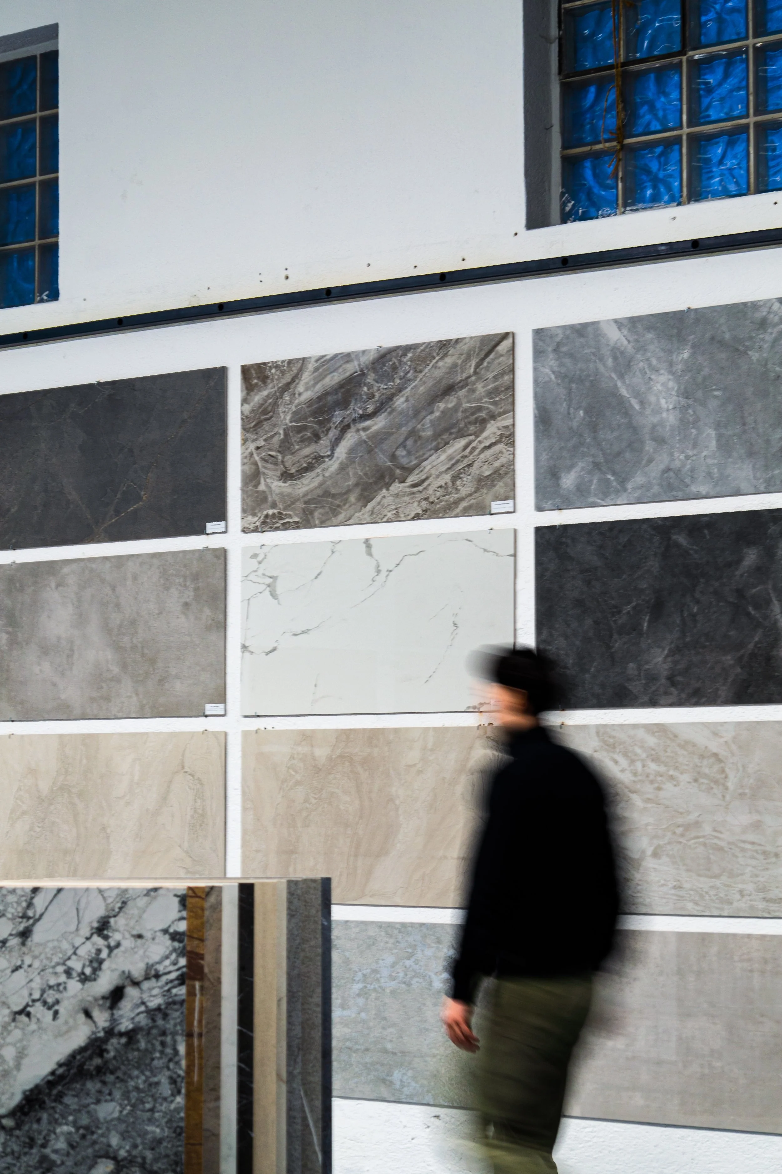 Ein Mensch läuft an einer Wand mit mehreren gefliesten Marmor- oder Steinfliesen vorbei, im Hintergrund sind zwei Fenster mit blauen Kunststoffscheiben sichtbar. Aufgenommen von Hilal Studios.