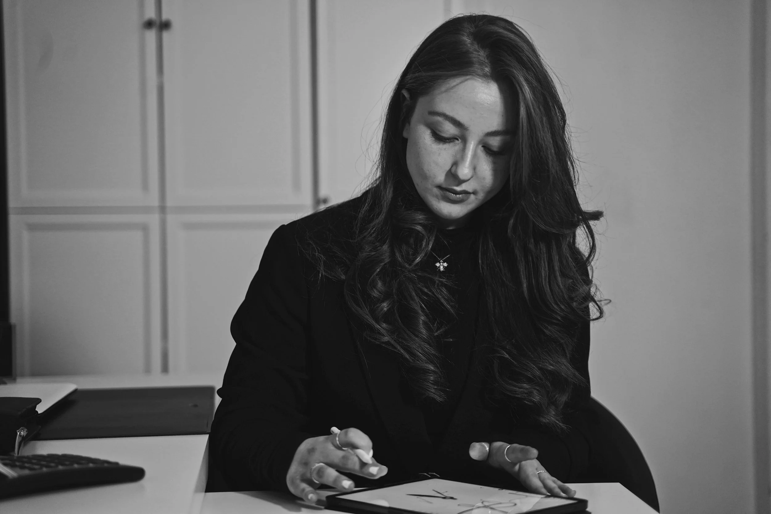 Schwarze und weiße Fotografie eines jungen Mädchens mit langen, gewellten Haaren, das an einem Tisch mit einem Zeichenblock oder Tablet arbeitet. Aufgenommen von Hilal Studios.