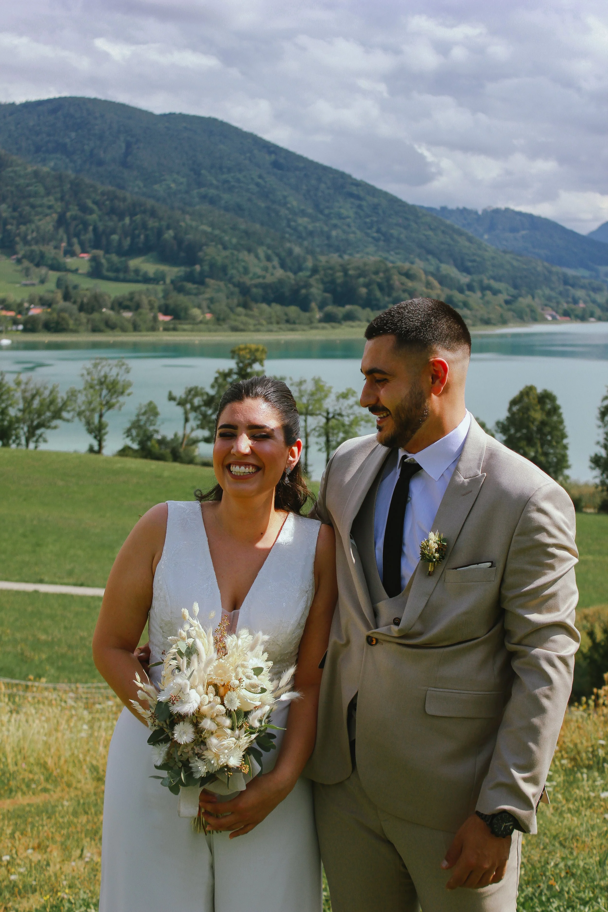 Ein lachendes Brautpaar bei einer Hochzeit im Freien mit einem See, Bergen und Wolken im Hintergrund. Aufgenommen von Hilal Studios.