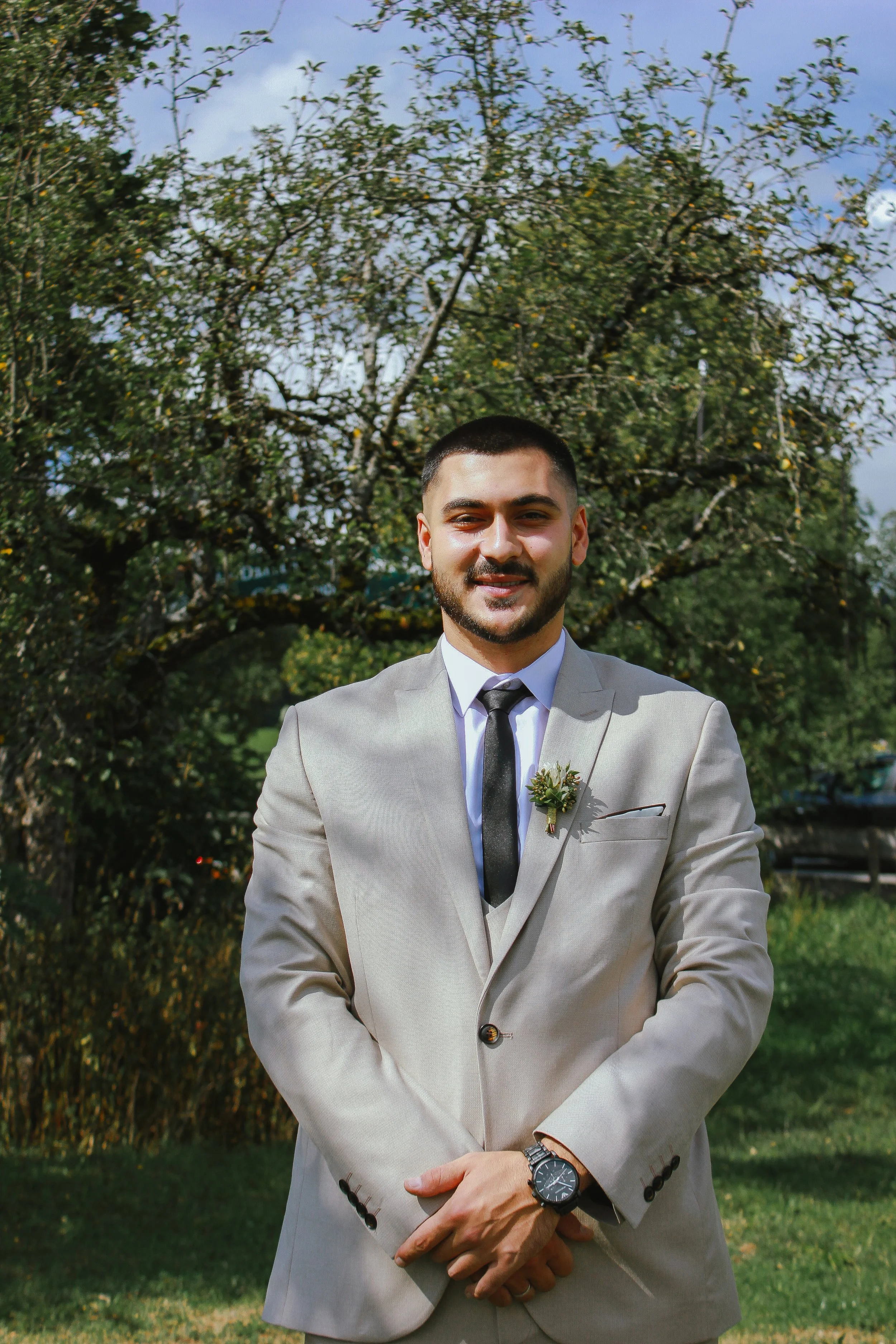 Junger Mann in einem hellgrauen Anzug mit Krawatte und Blumenansteckdose, stehend im Freien vor einem Baum. Aufgenommen von Hilal Studios.