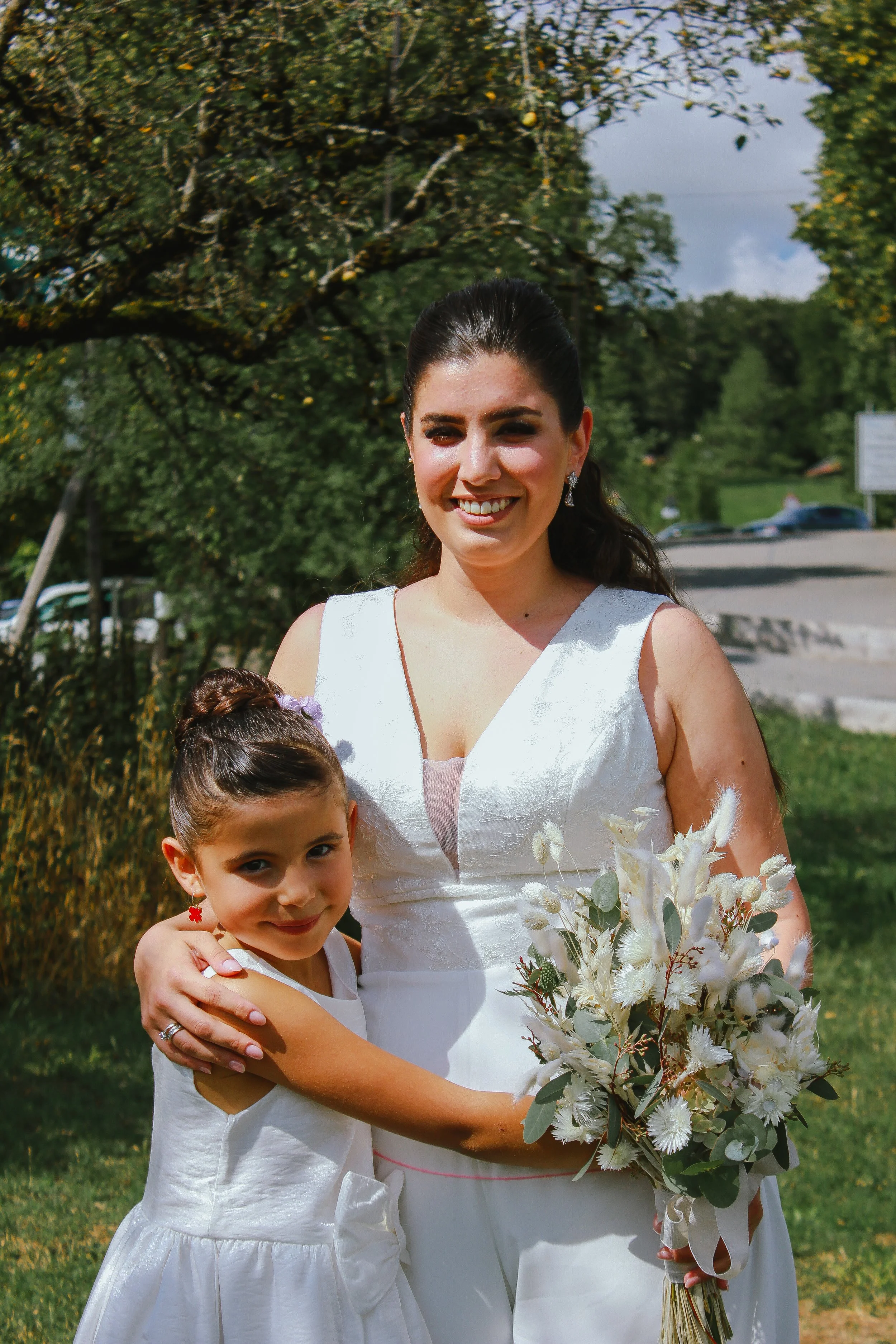 Eine Frau in einem weißen Brautkleid hält ein Blumenbouquet, während ein junges Mädchen sie umarmt und beide lächeln. Es ist ein sonniger Tag im Freien, mit Bäumen im Hintergrund. Aufgenommen von Hilal Studios.