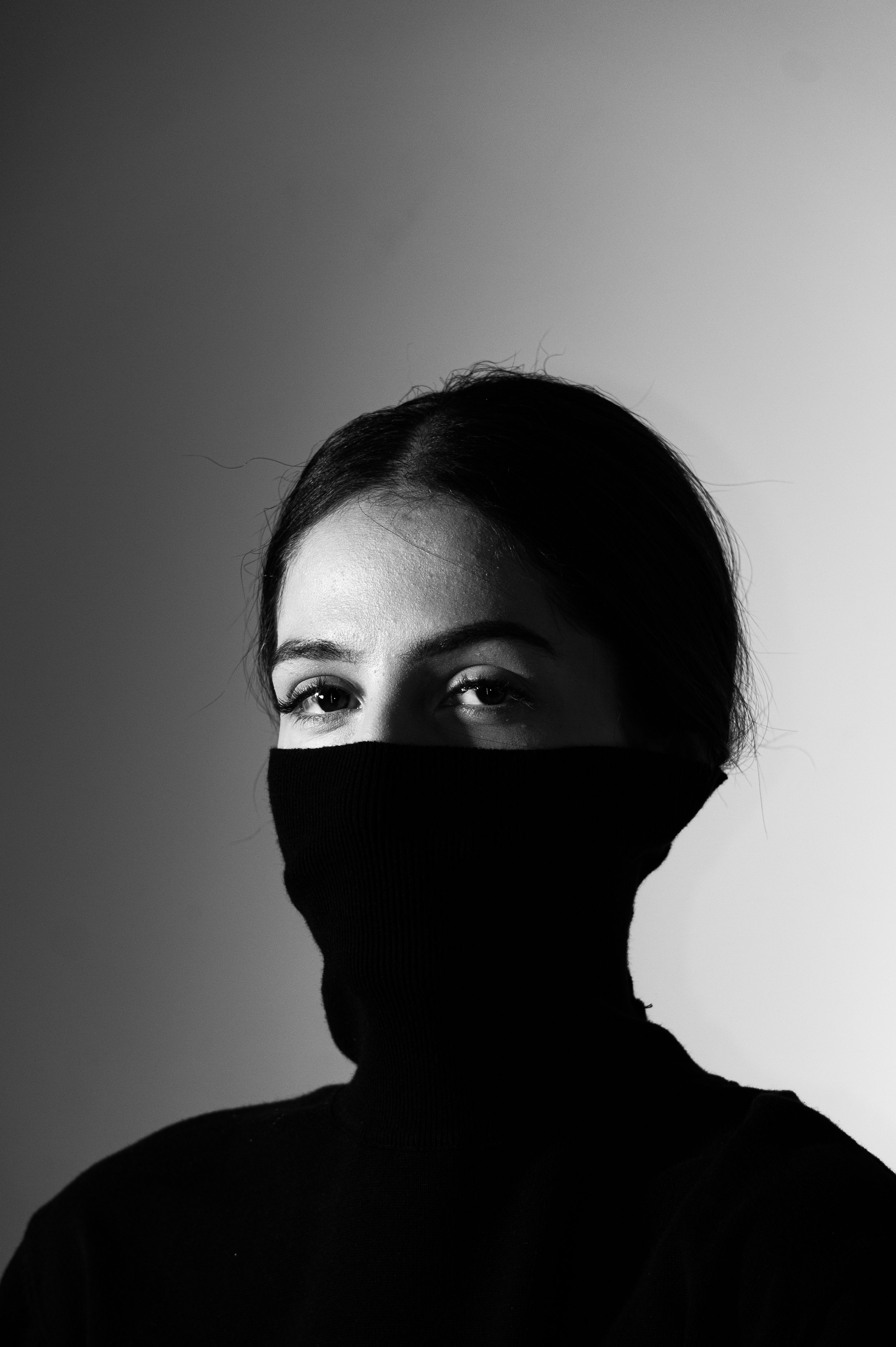 Schwarz-Weiß-Foto einer jungen Frau mit schwarzen Haaren und schwarzer Gesichtsmaske, die nur ihre Augen sichtbar lässt, vor einem zweifarbigen Hintergrund. Aufgenommen von Hilal Studios.