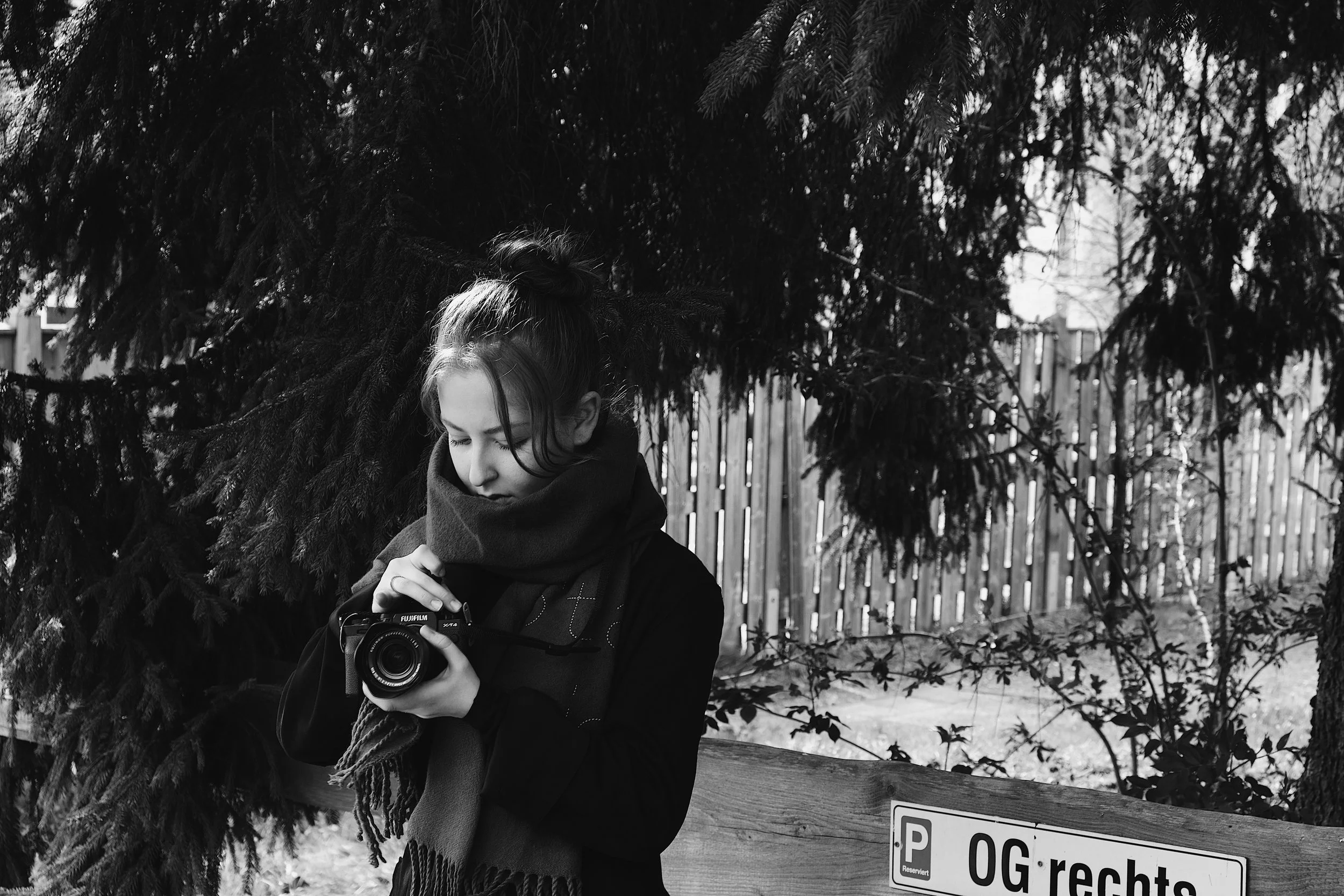 Junge Frau mit dunklem Mantel und Schal fotografiert mit einer Digitalkamera, steht vor einem Baum und Zaun, in schwarz-weiß. Aufgenommen von Hilal Studios.