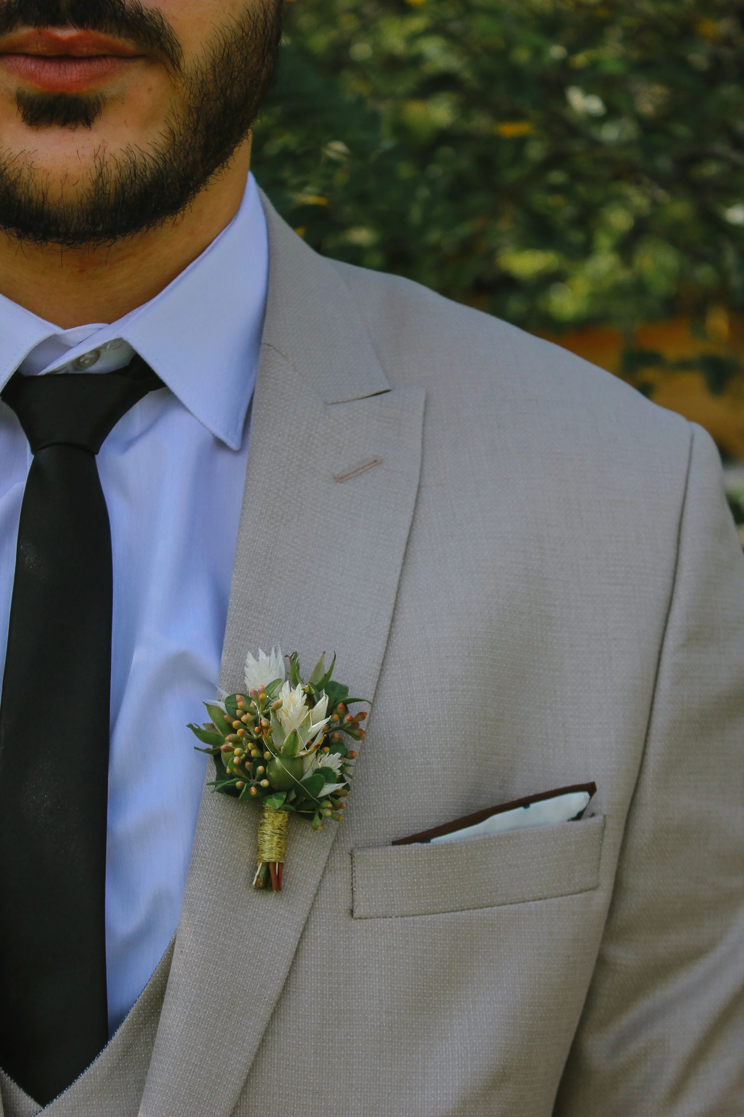 Nahaufnahme eines Mannes in einem hellgrauen Anzug, weißem Hemd, schwarzer Krawatte, mit Boutonnière und einstecktem Taschentuch in der Brusttasche. Aufgenommen von Hilal Studios.