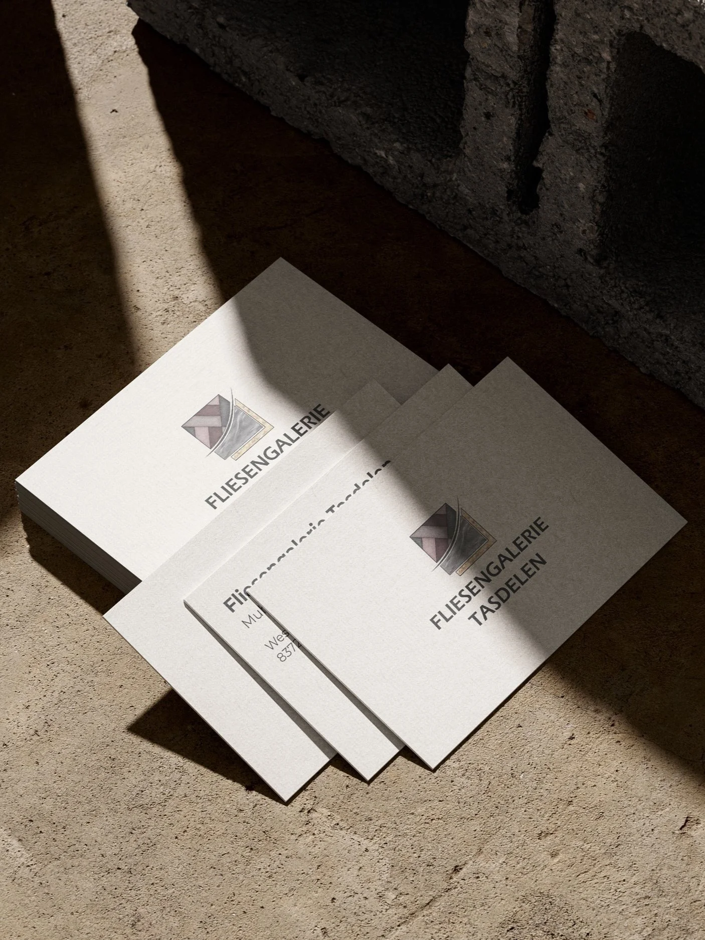 Drei weiße Visitenkarten auf einem Sandboden, mit Schatten und einem Steinfundament im Hintergrund, von der Firma Fliesengalerie Taschen. Erstellt von Hilal Studios.