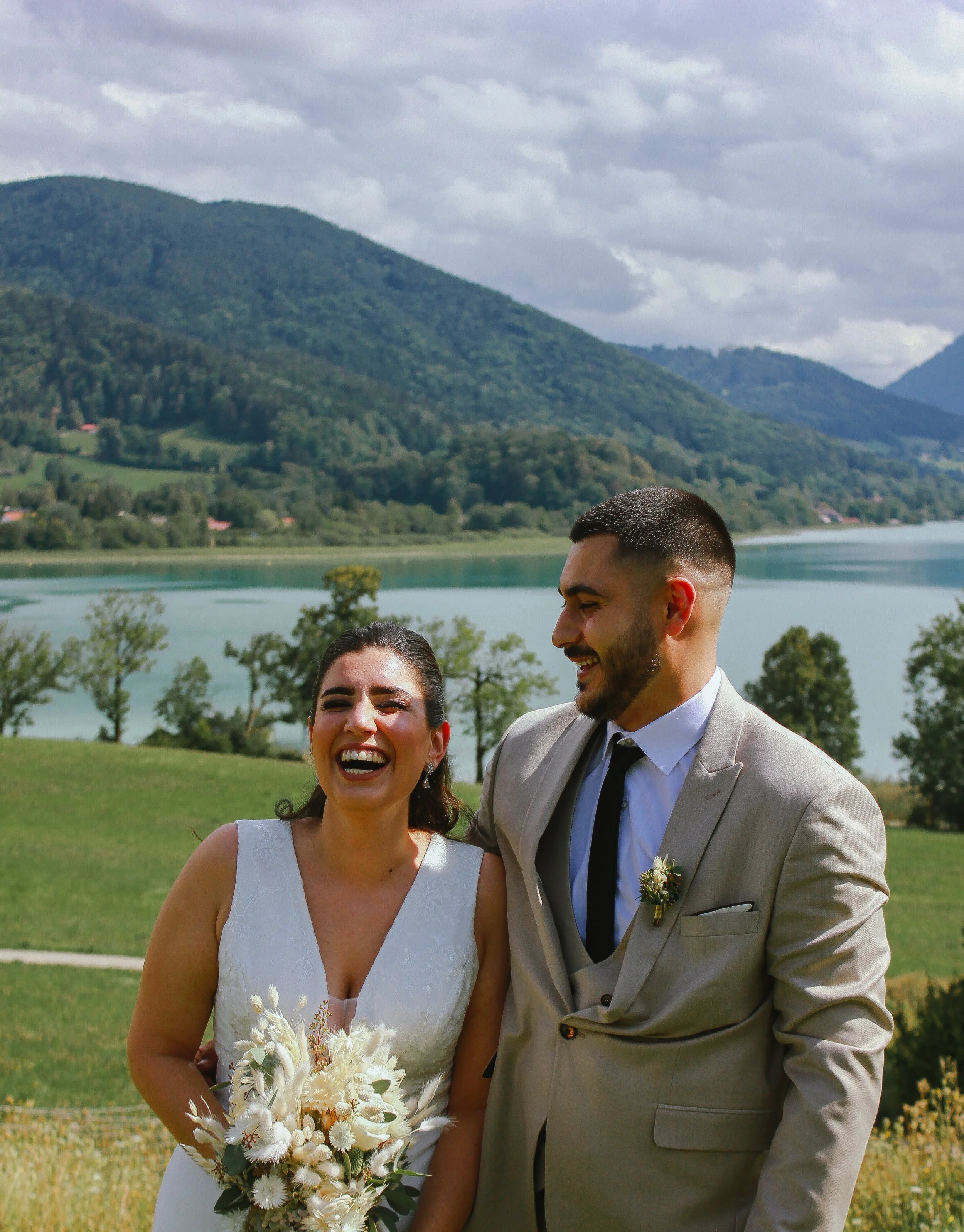 Ein lachendes Brautpaar bei einer Hochzeit im Freien vor einer malerischen Landschaft mit Bergen, Wasser und Bäumen. Aufgenommen von Hilal Studios.