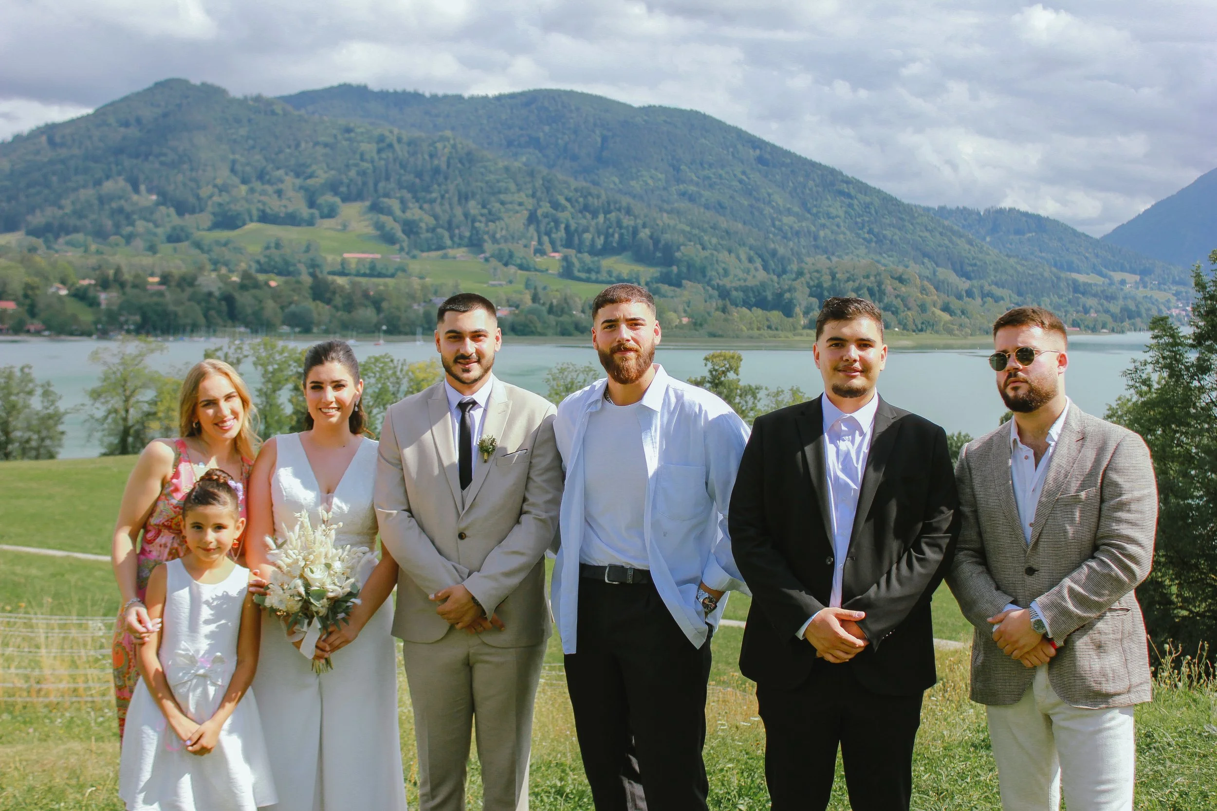 Gruppe von sieben Menschen in formeller Kleidung, vor einer Seen- und Berglandschaft, bei einer Feier im Freien. Aufgenommen von Hilal Studios.